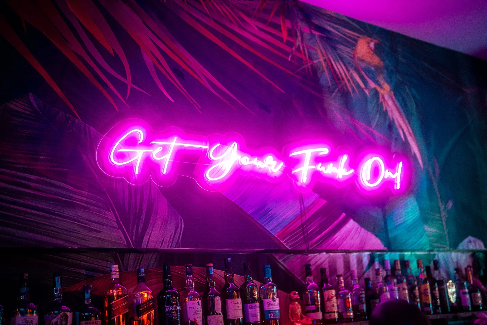 Neon sign in pink lights saying "Get Your F*** On" behind a bar with a variety of liquor bottles.