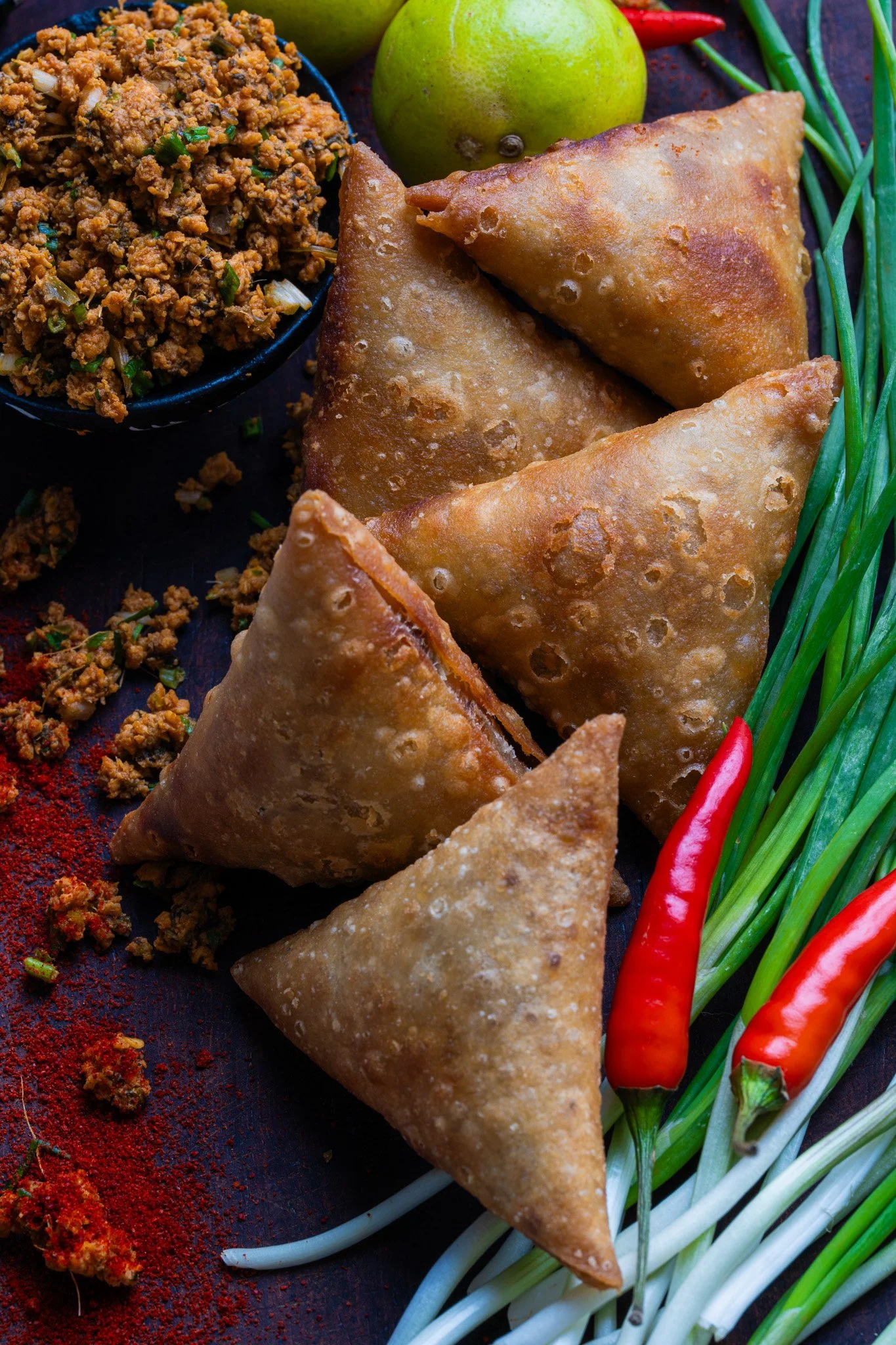 Samosas arranged on a black platter with green onions and red chili peppers on the side, alongside a small bowl of seasoned ground meat, with a lime and green chilies in the background.