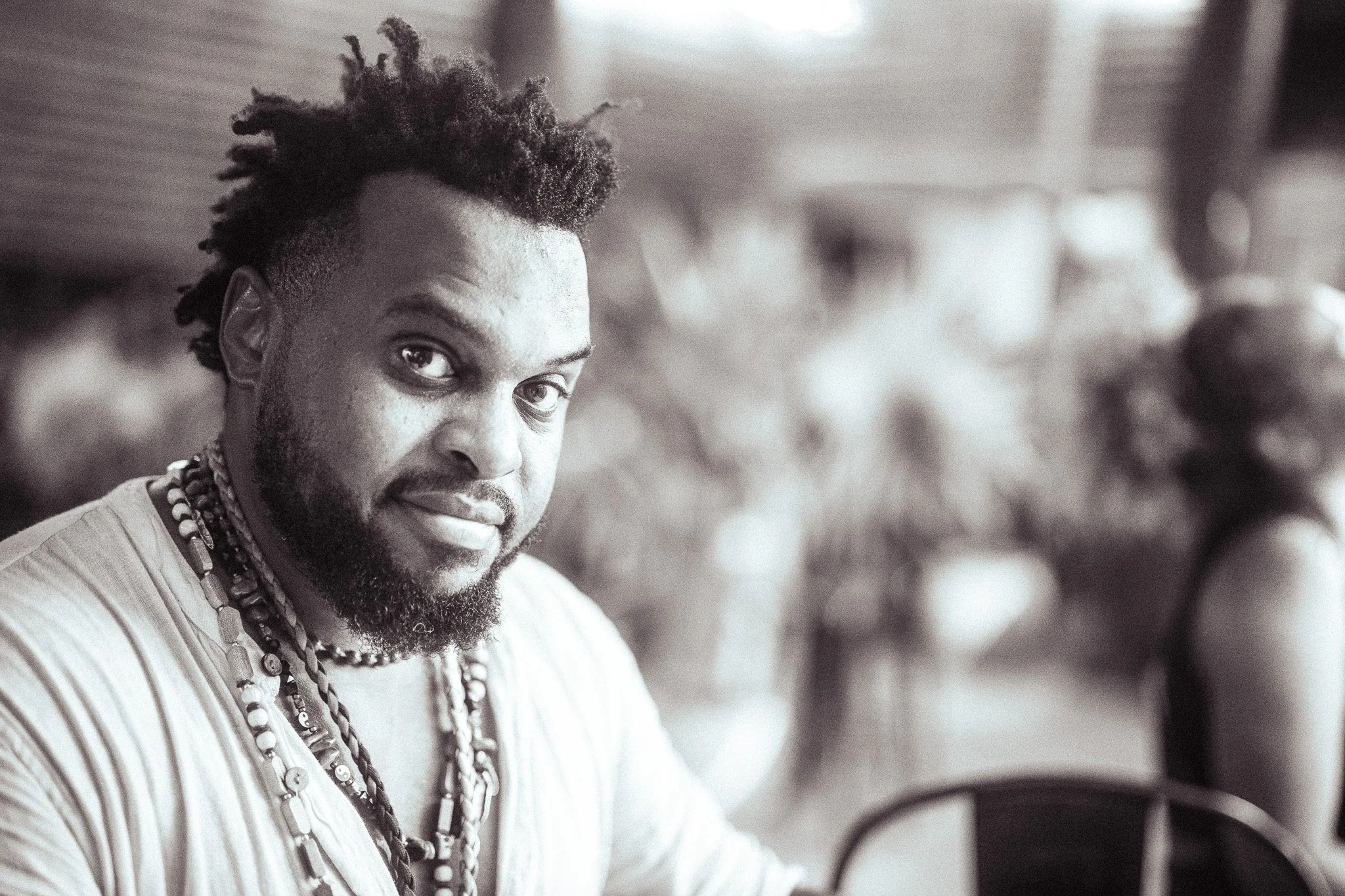 A Black man with a beard and dreadlocks wearing layered necklaces and a light-colored shirt, looking at the camera with a slight smile, in a setting that appears to be a market or outdoor area.