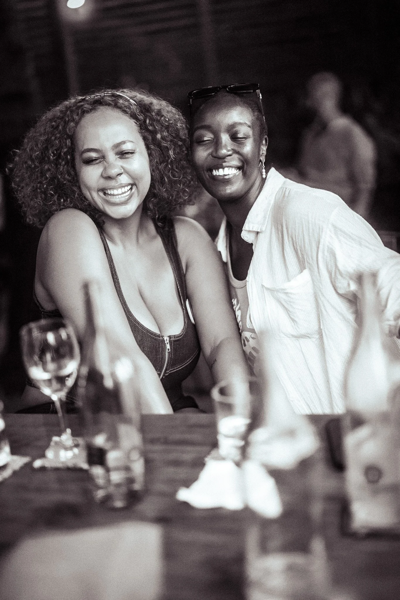 Two women smiling and laughing together at a social gathering, with drinks and bottles on the table.