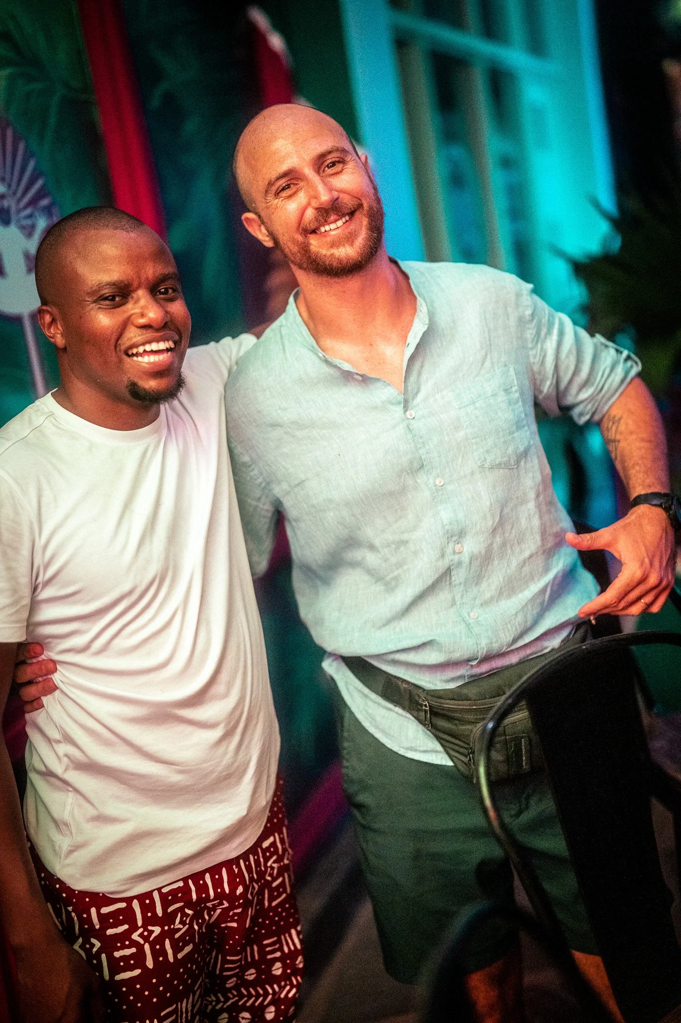 Two men smiling and standing close together indoors, with one man wearing a white T-shirt and patterned shorts, and the other in a light blue button-up shirt and dark shorts, in a colorful setting.