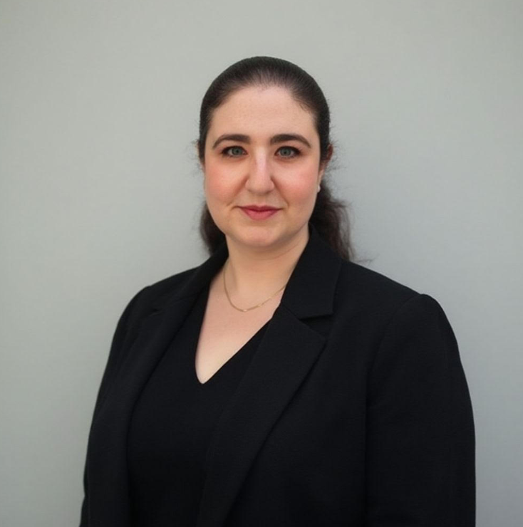 Portrait of a woman with dark brown hair in a ponytail, wearing a black blazer and top, standing against a plain light gray background.