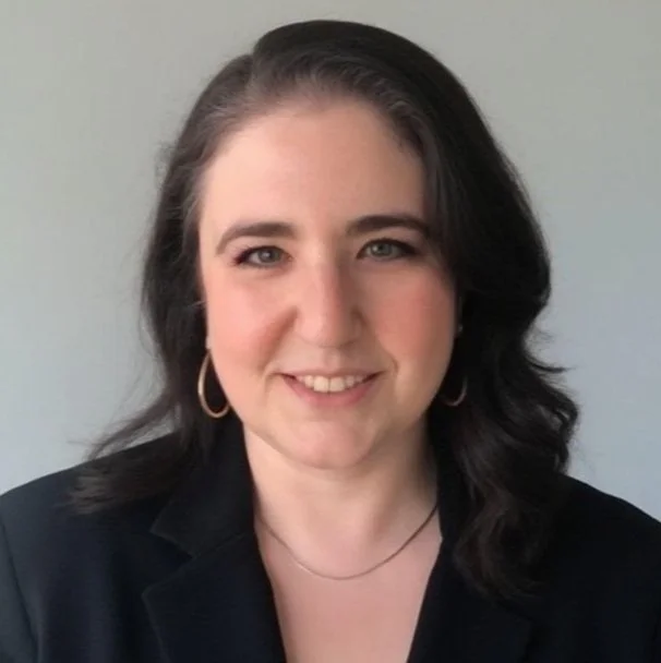 A woman with shoulder-length dark hair, light skin, and a friendly smile, wearing gold hoop earrings, a black blazer, and a thin necklace, standing against a plain light-colored background.