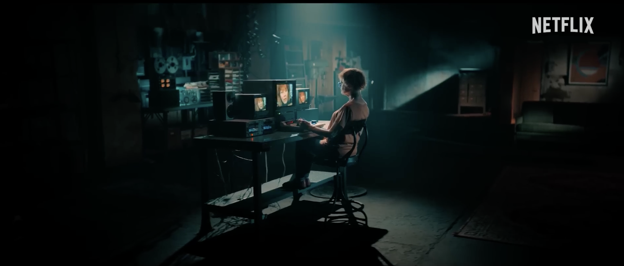 A woman sits at a desk with multiple monitors displaying faces and scenes, in a dimly lit room with vintage electronic equipment and bookshelves, illuminated by a soft blue light.