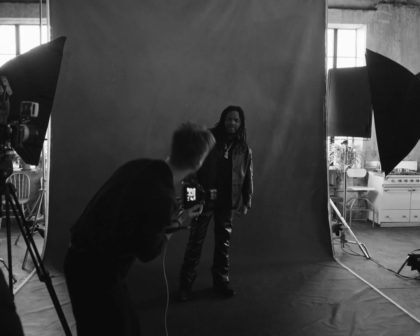 A person with dreadlocks and a leather jacket is standing in front of a photography backdrop, being photographed by another person holding a camera. Studio lights are set up on either side of the backdrop, and a kitchen is visible in the background.