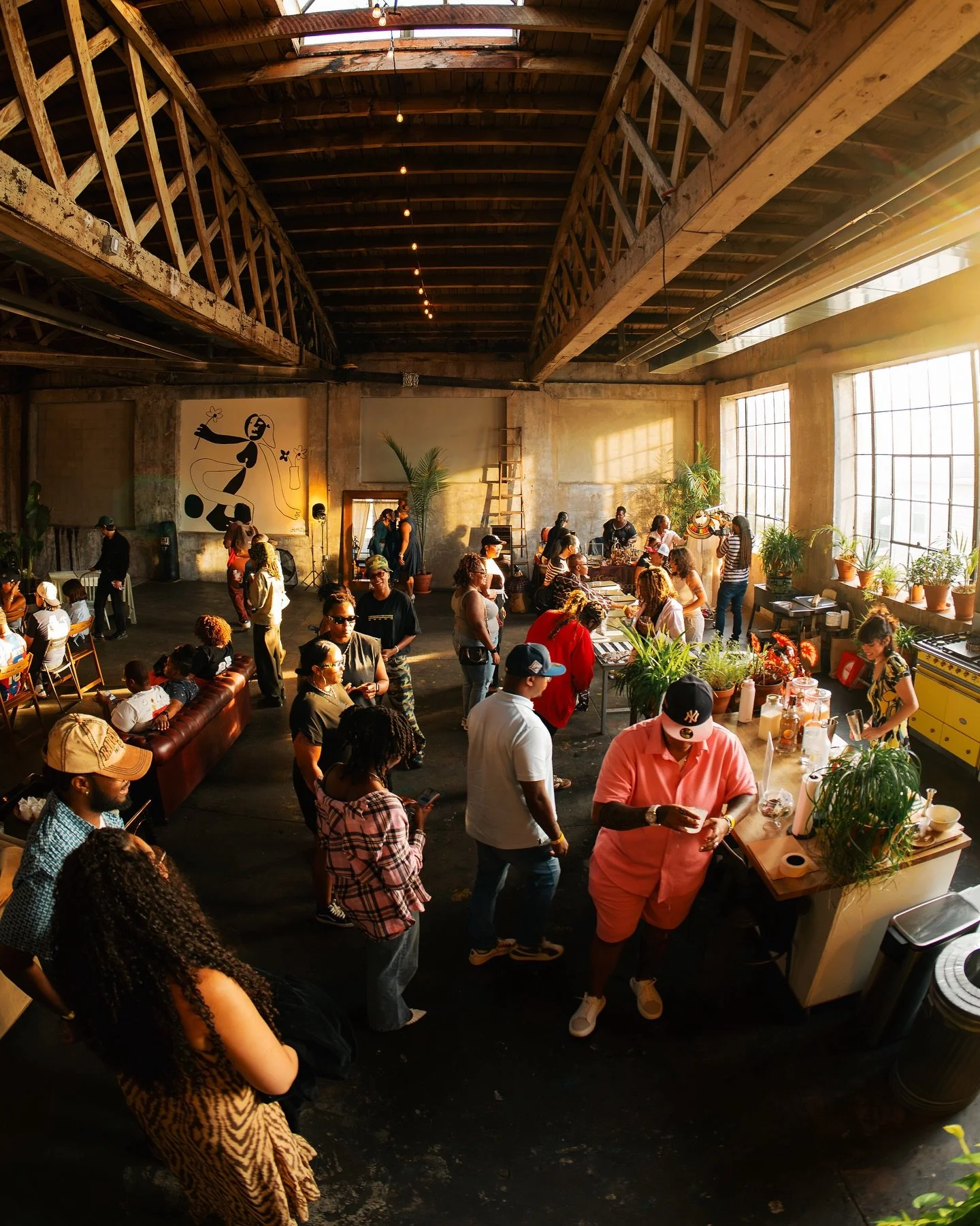 A lively indoor gathering with people socializing, some standing and some seated, in a spacious, industrial-style venue with large windows, potted plants, and artwork on the walls. The sun is shining through the windows, illuminating the room.