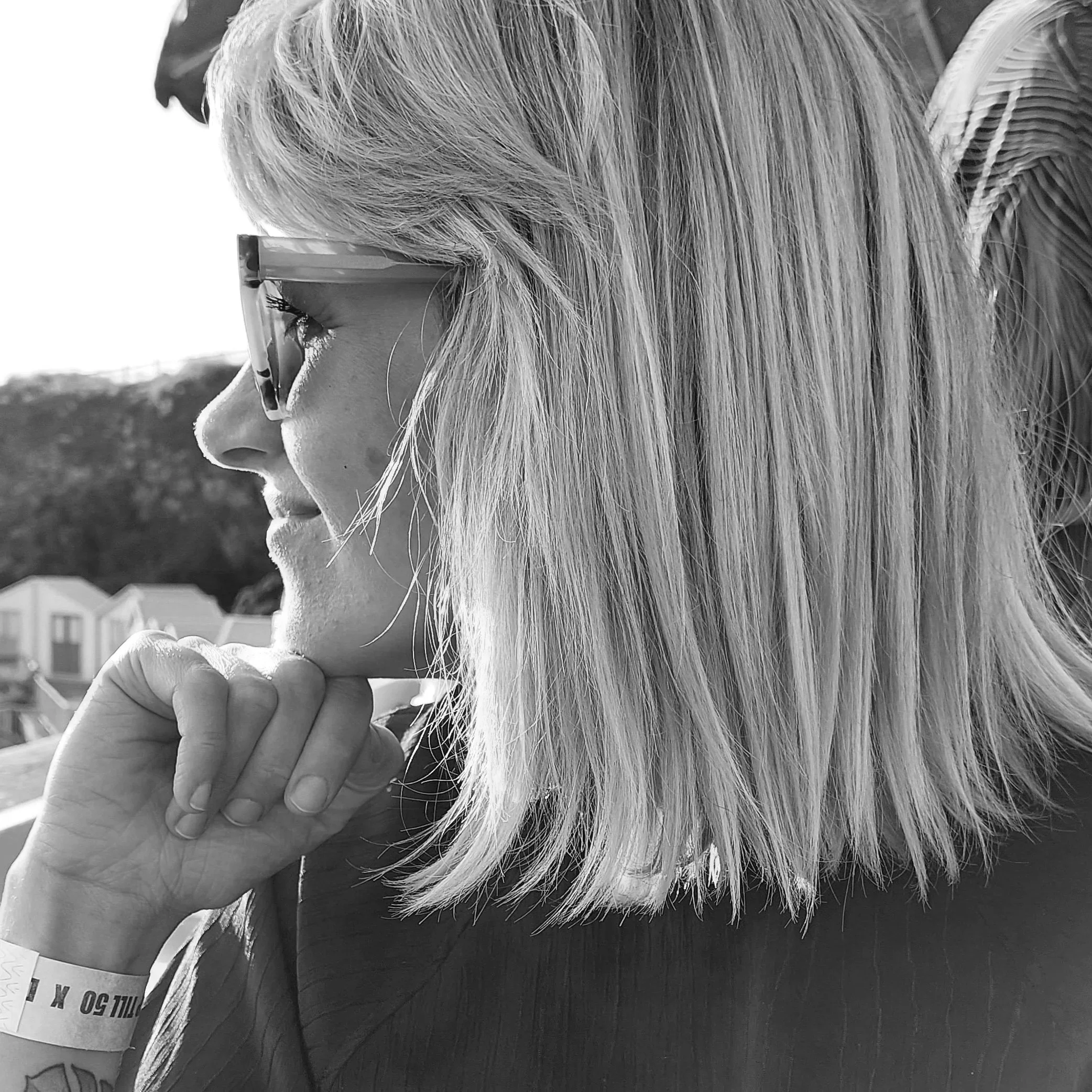 A black and white photo of a woman with straight, shoulder-length hair and glasses, looking sideways with her chin resting on her hand. Houses and hills are visible in the background.