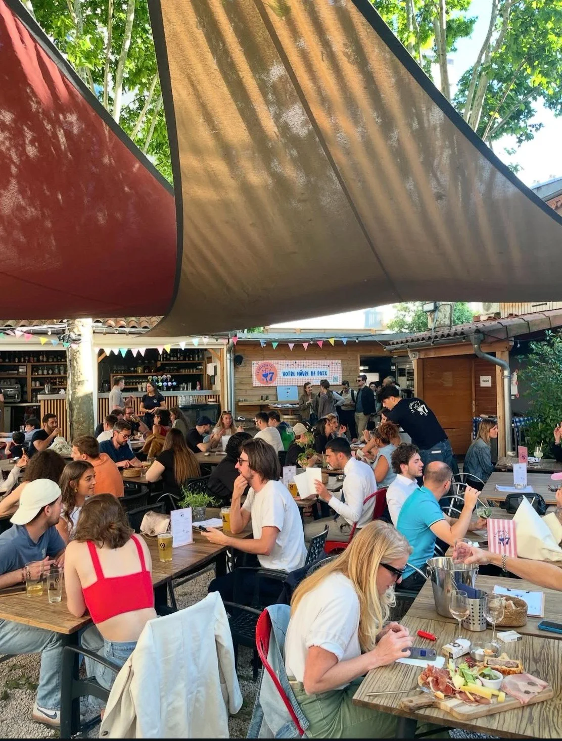 Une grande terrasse extérieure avec de nombreuses personnes assises à des tables, mangeant et discutant. Il y a une structure en toile colorée au-dessus pour l'ombre.
