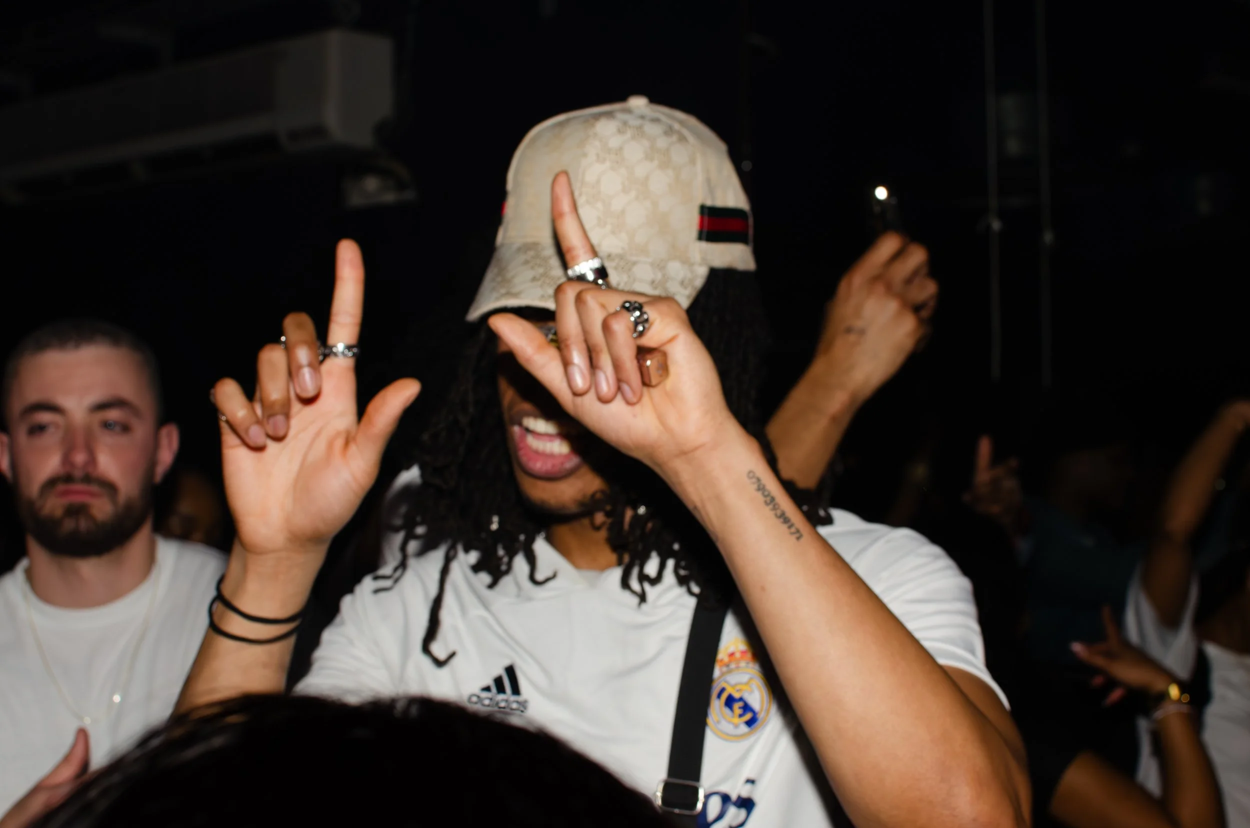 People dancing in a dimly lit club with one person wearing a cap and making a hand gesture.