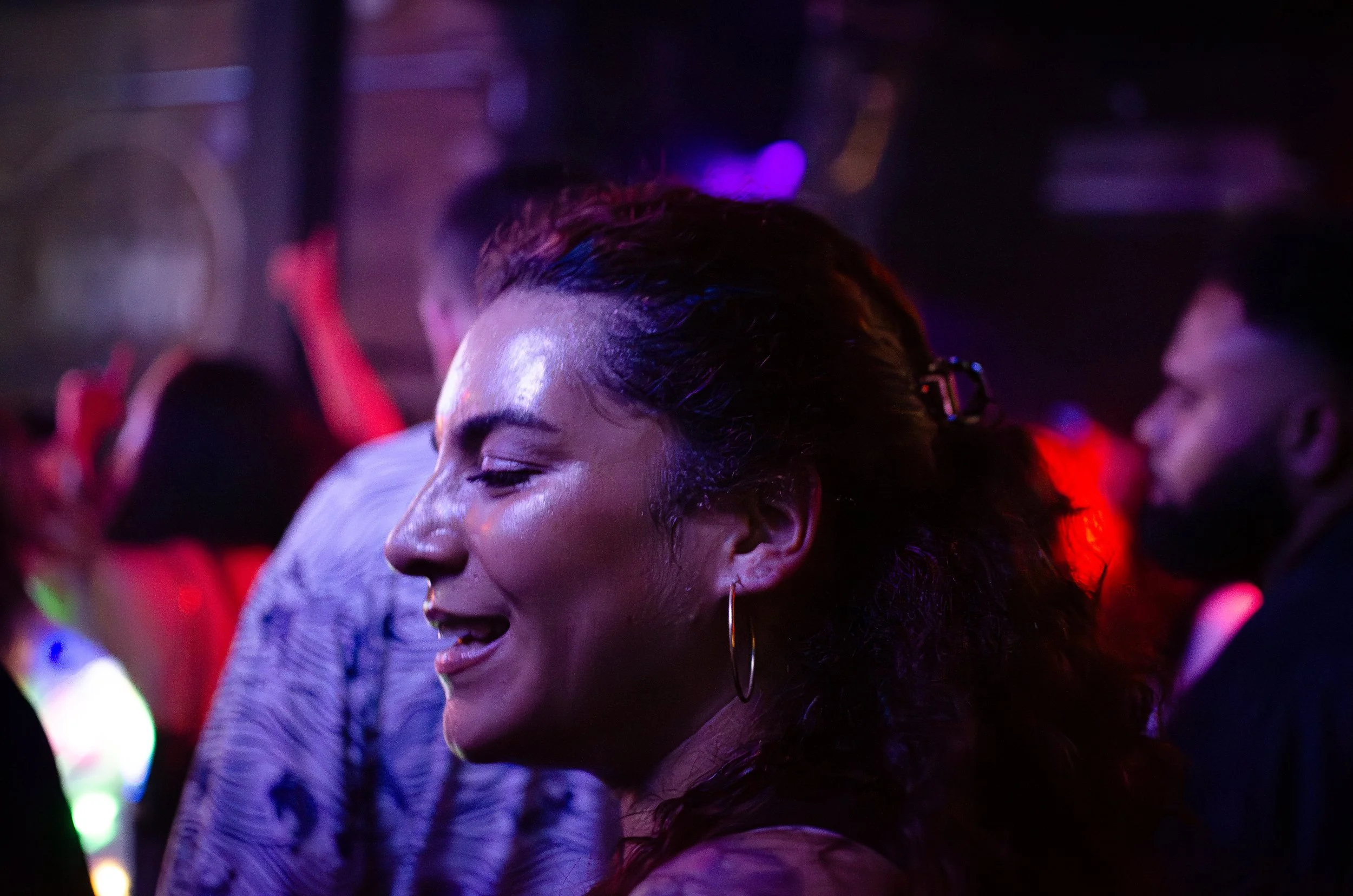 Woman with hoop earrings smiling in a dimly lit, crowded nightclub.