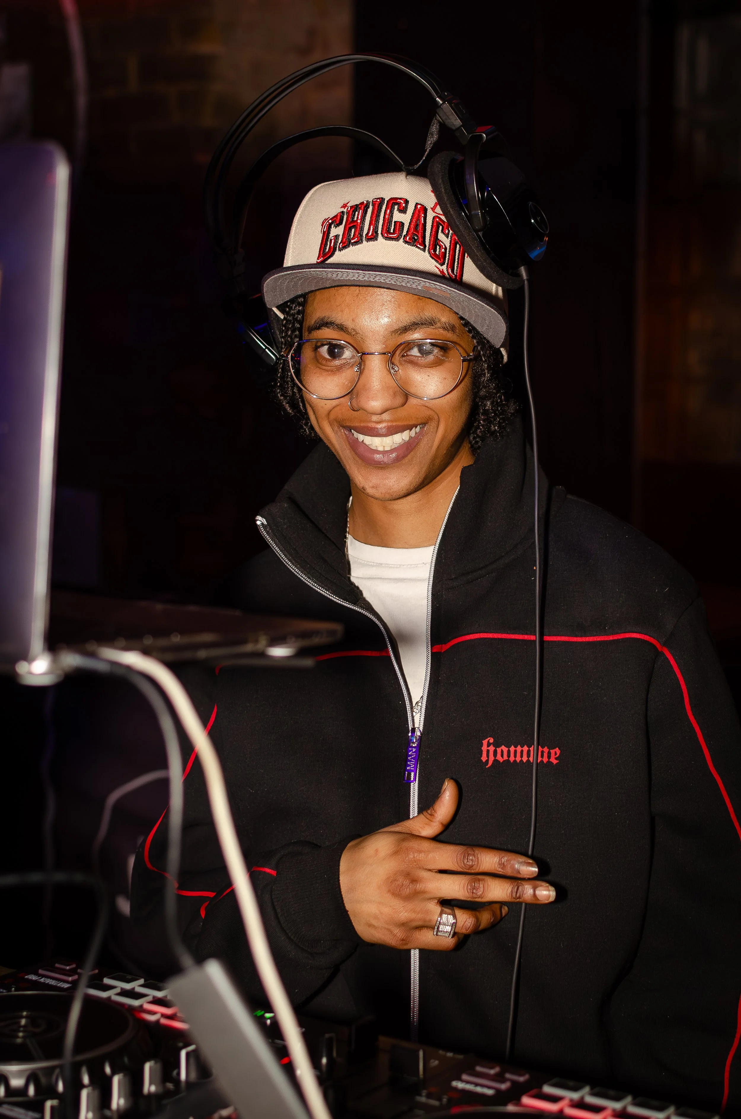 Person in a "Chicago" cap with headphones, smiling, operating DJ equipment.