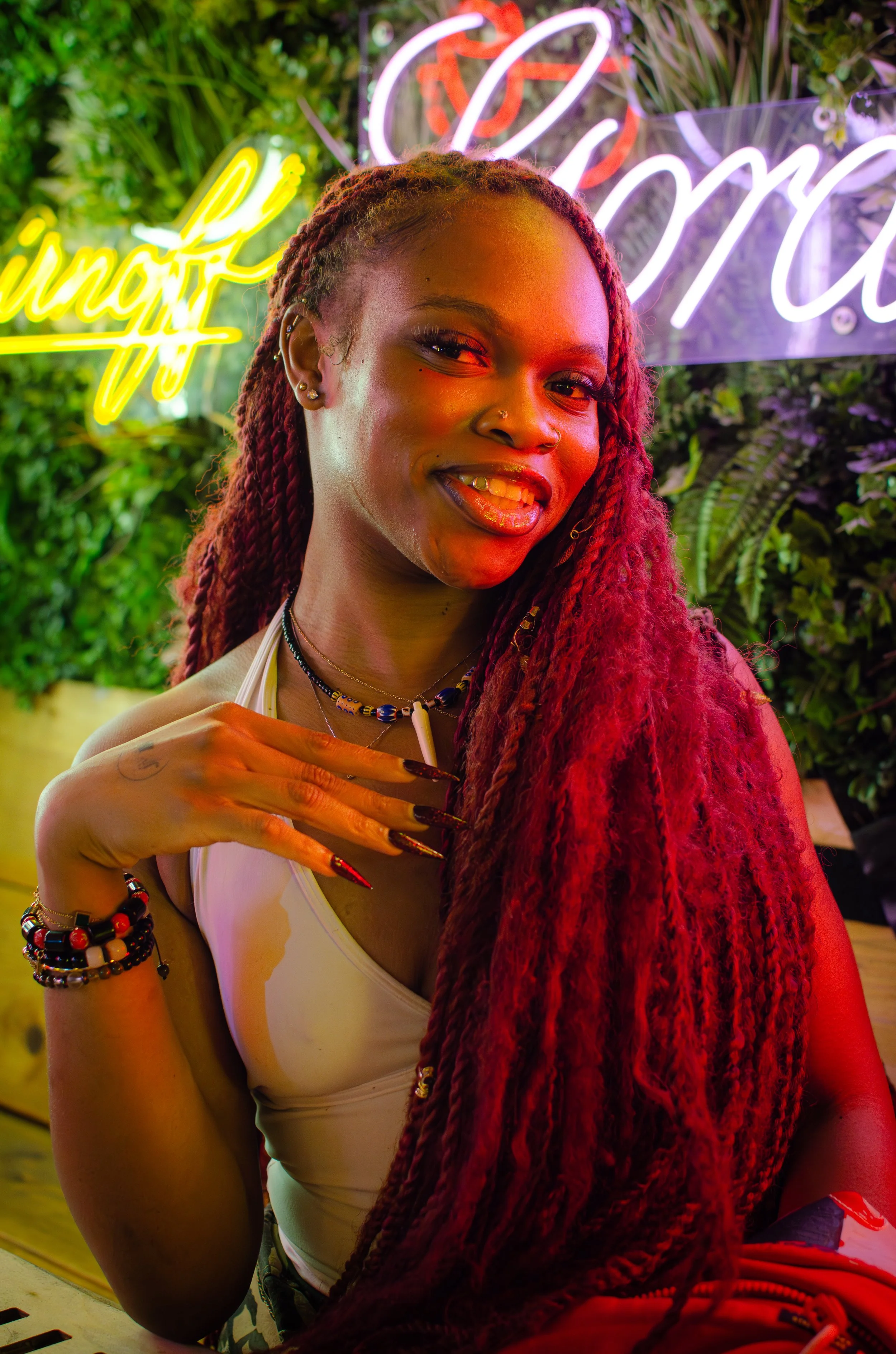 Person with long red braids smiling in front of neon lights and greenery background.
