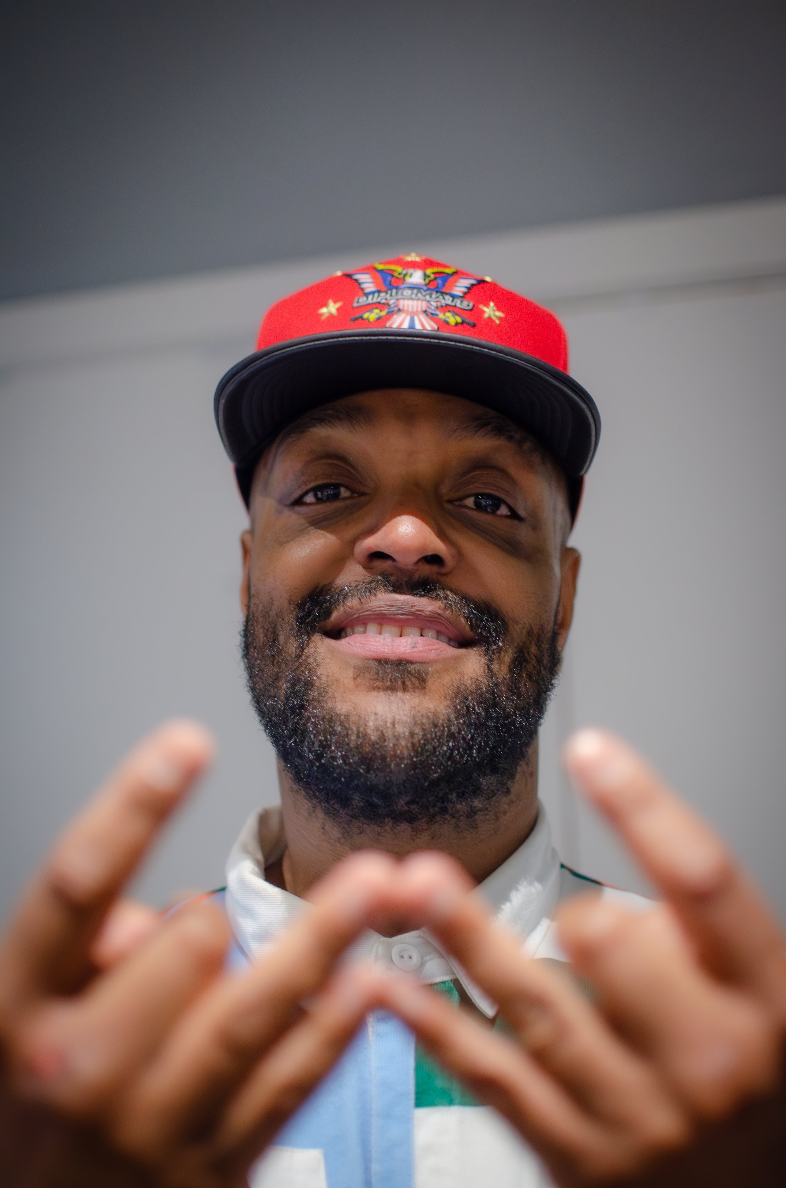 Man wearing a red hat with emblem, smiling, and holding his hands in a gesture in front of him.