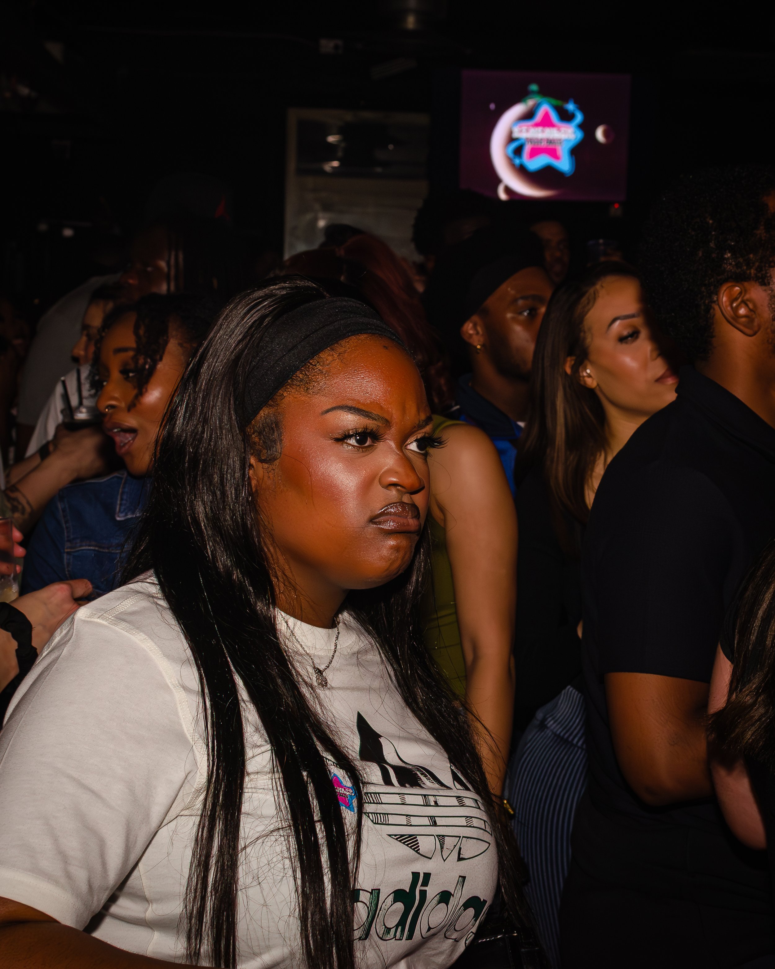 A woman in a white t-shirt with a graphic, standing in a crowded indoor setting, appears to be making a displeased expression. Other people are visible in the background, with a screen displaying a logo.