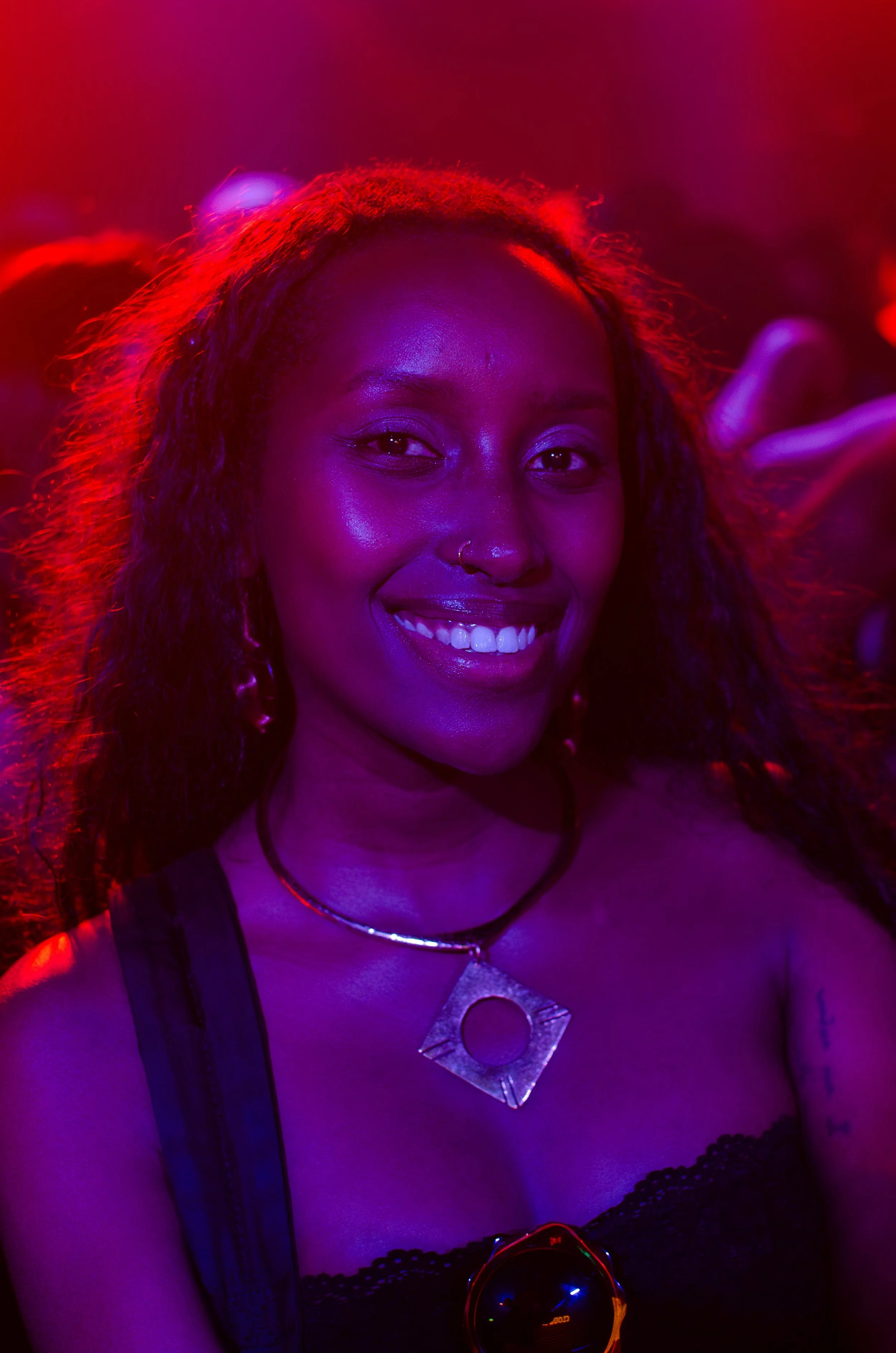 Smiling woman with long hair, wearing a necklace and strapless top, in a nightclub setting with red and purple lighting.