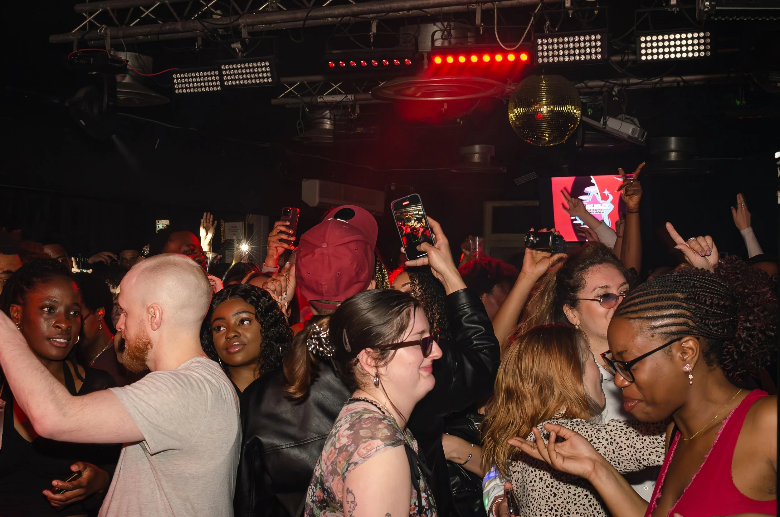 People dancing and taking photos in a crowded nightclub with red lighting and a disco ball.