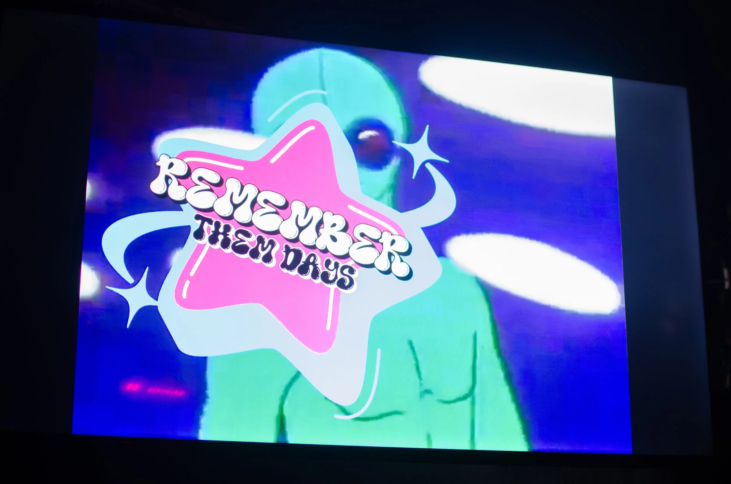 A digital image featuring a green alien figure on a large screen with colorful, distorted effects. In front, there is a pink star with the text "Remember Them Days" in a retro-style font. The background is a mix of purple and blue colors with white o