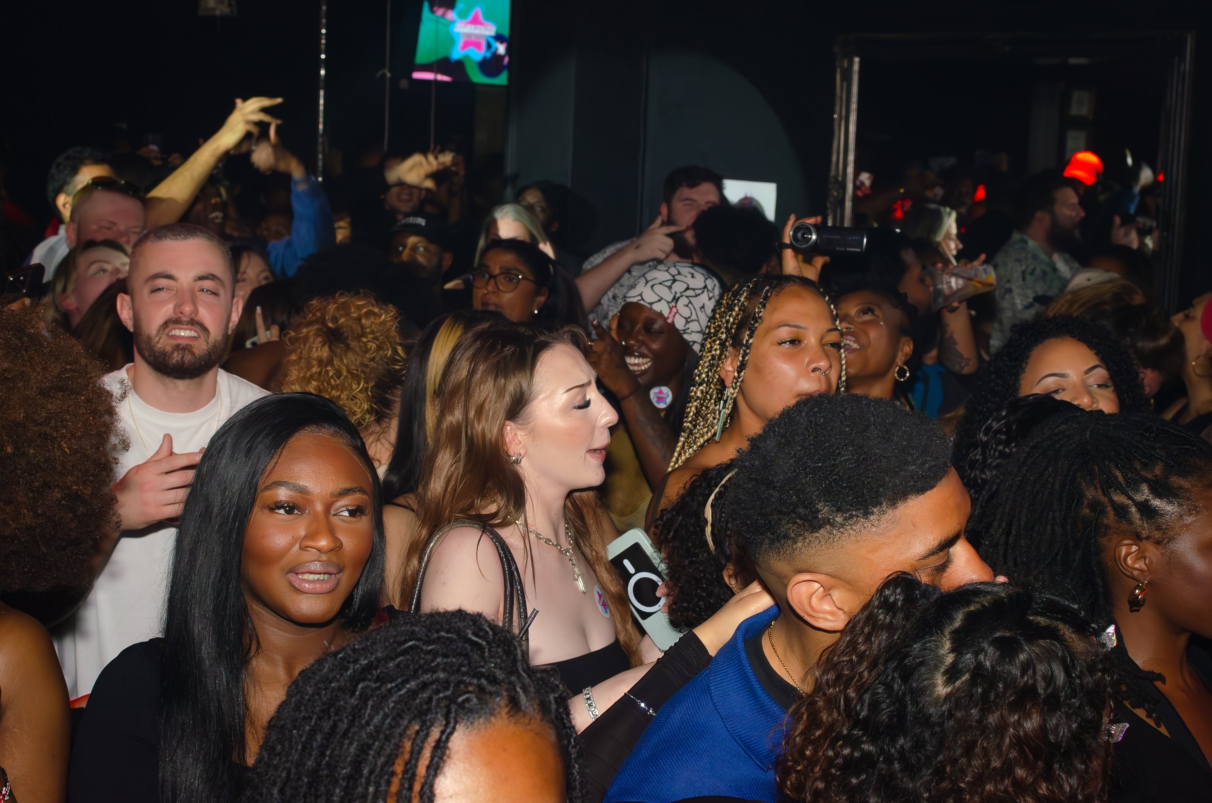 A crowded group of people in a lively nightclub setting, some smiling and interacting, with dim lighting and a dark background featuring colorful lights.