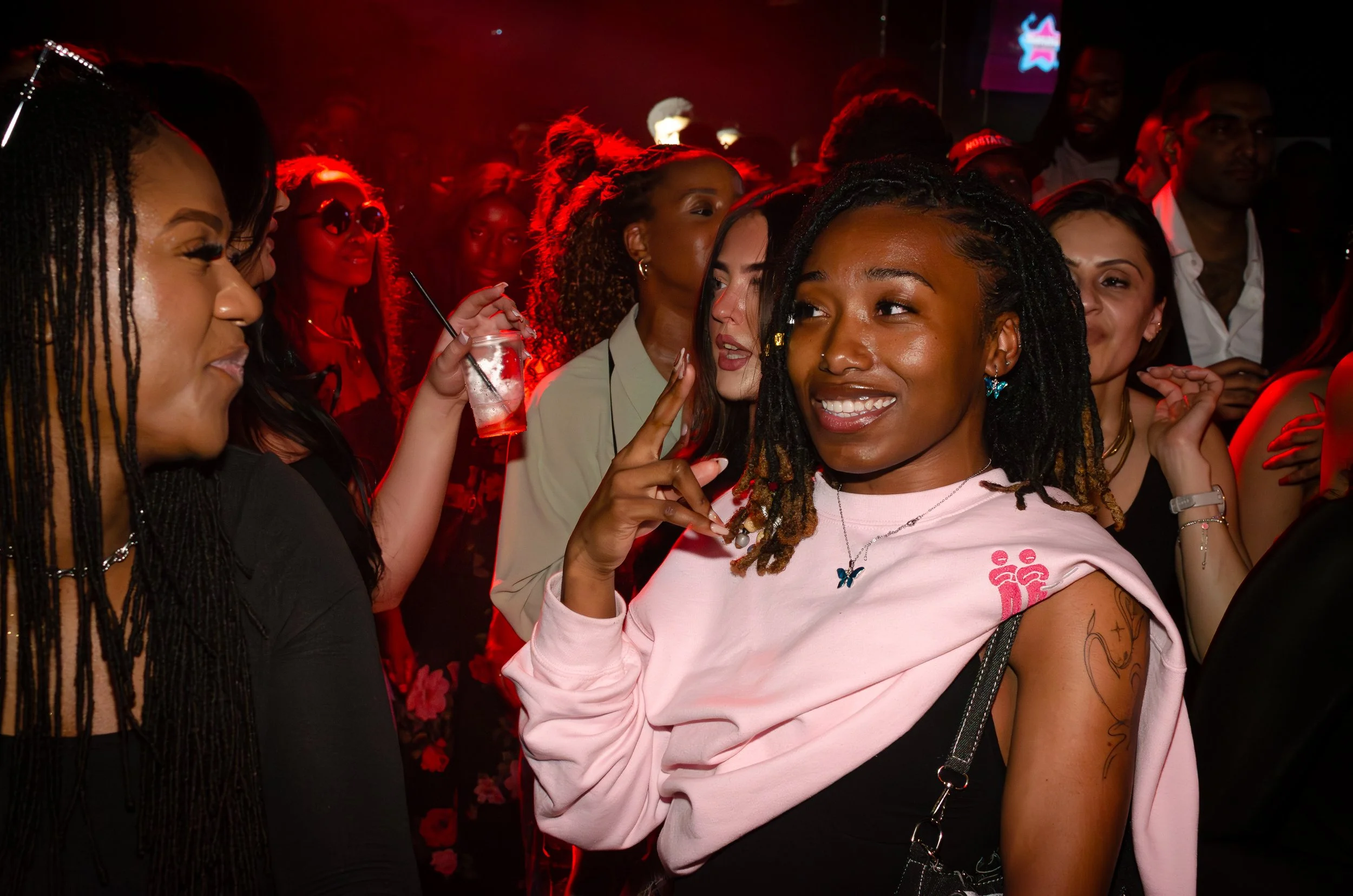 Group of people at a party with red lighting; a smiling person in a pink shirt is in the foreground.