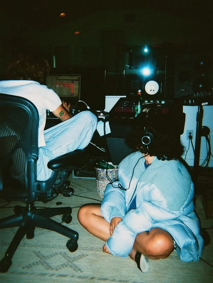 Two people in a recording studio, one sitting at a computer and the other sitting on the floor with headphones, both wearing casual clothes and in a dimly lit room with various equipment.
