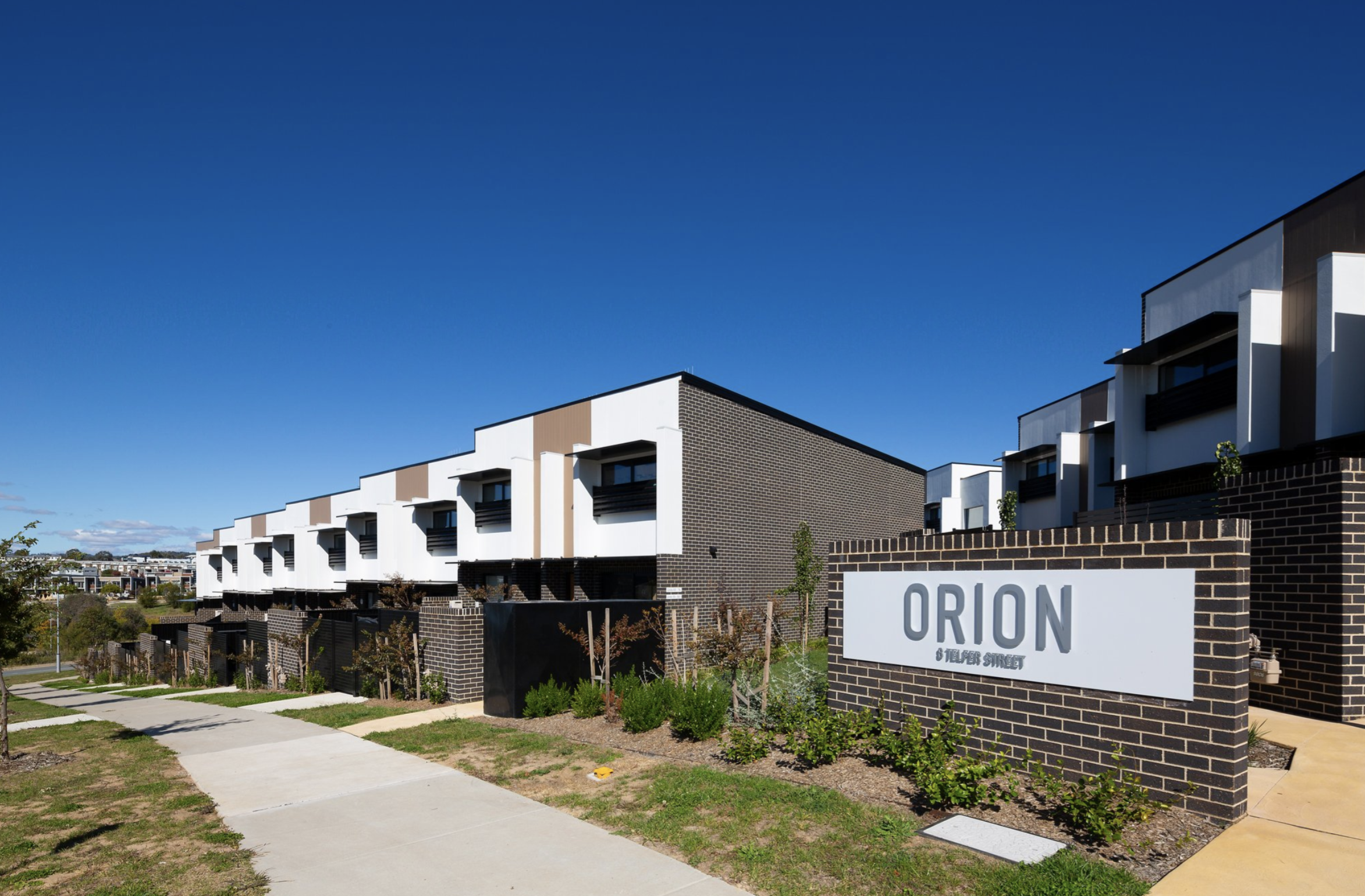 Modern townhouse complex named Orion with brown brick and white exterior, clear blue sky background.