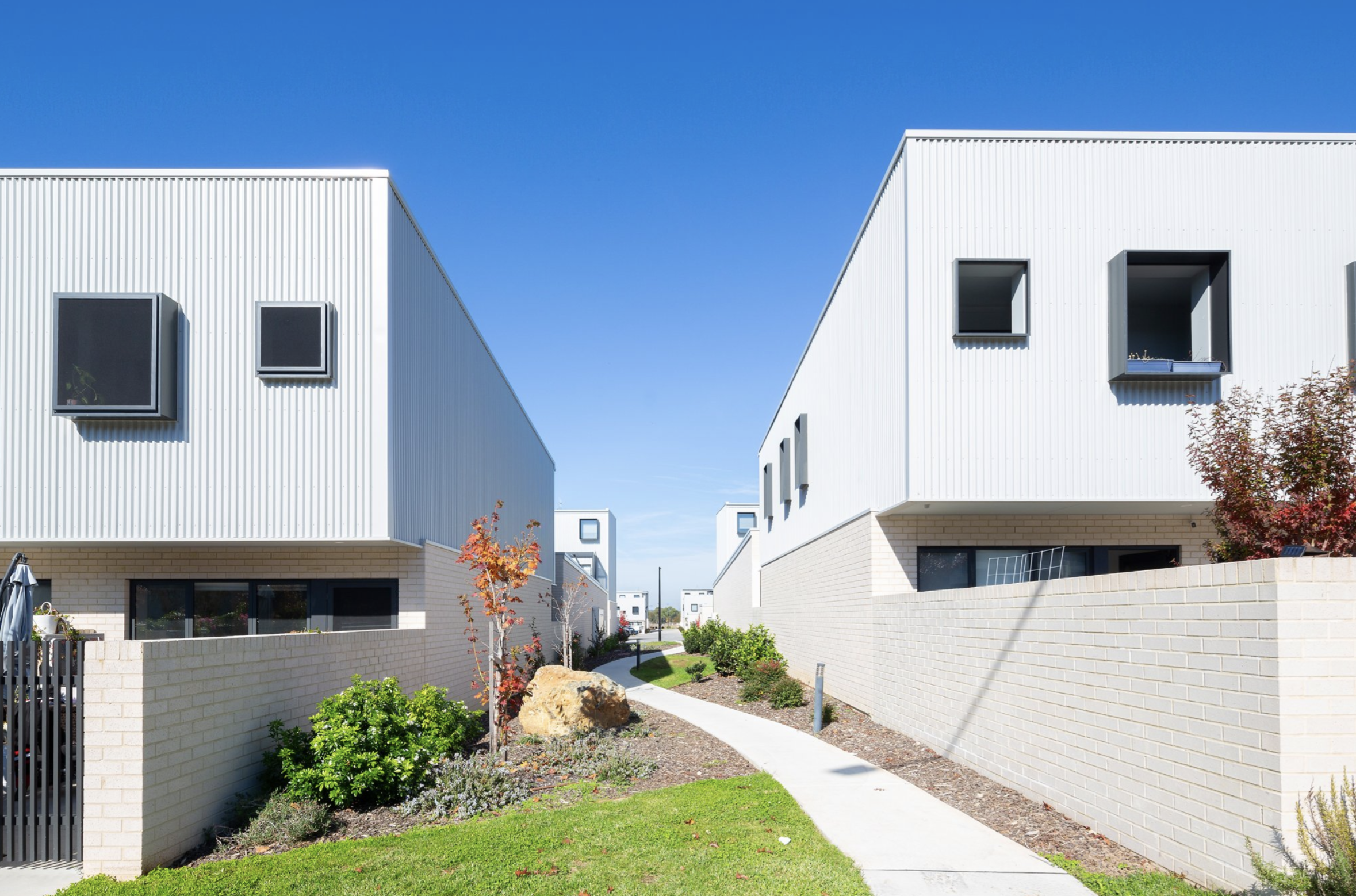 Modern residential buildings with flat roofs and white walls, separated by a narrow path with green landscaping under a clear blue sky.