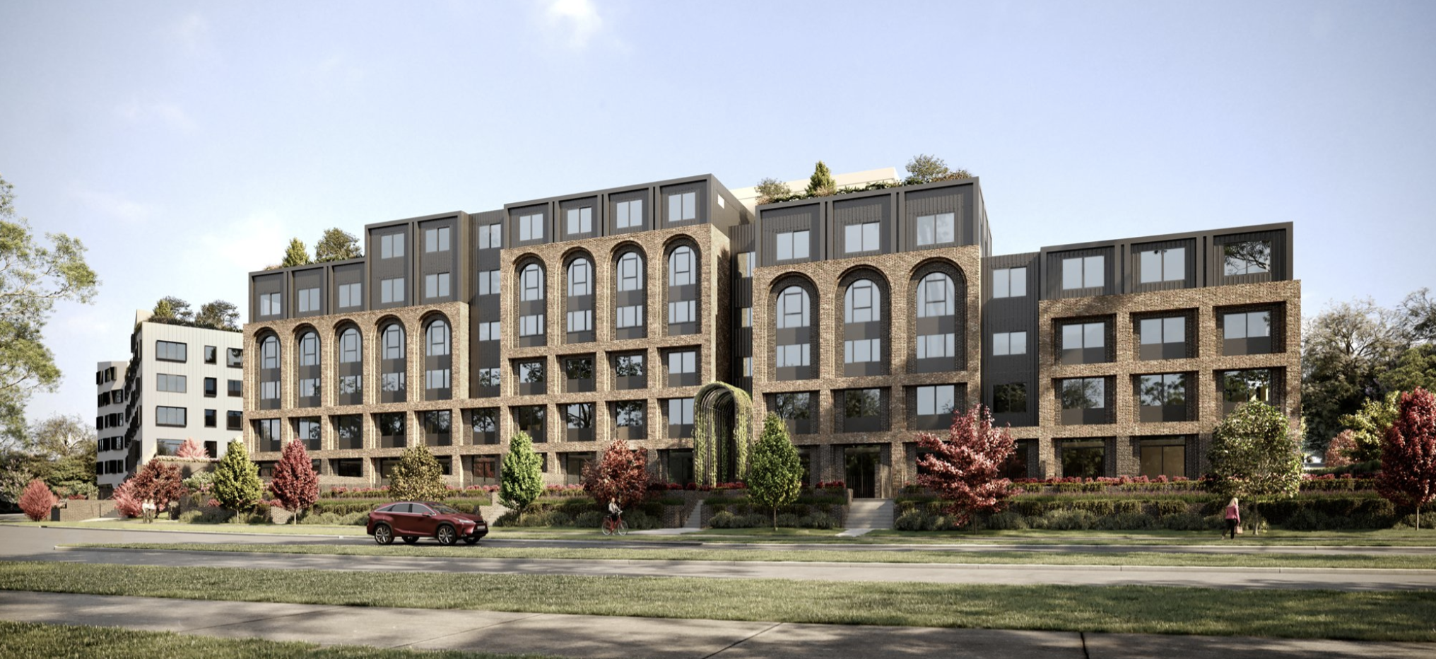 Modern apartment building with large windows and brick facade, surrounded by greenery and trees, with a car and cyclist on the road.