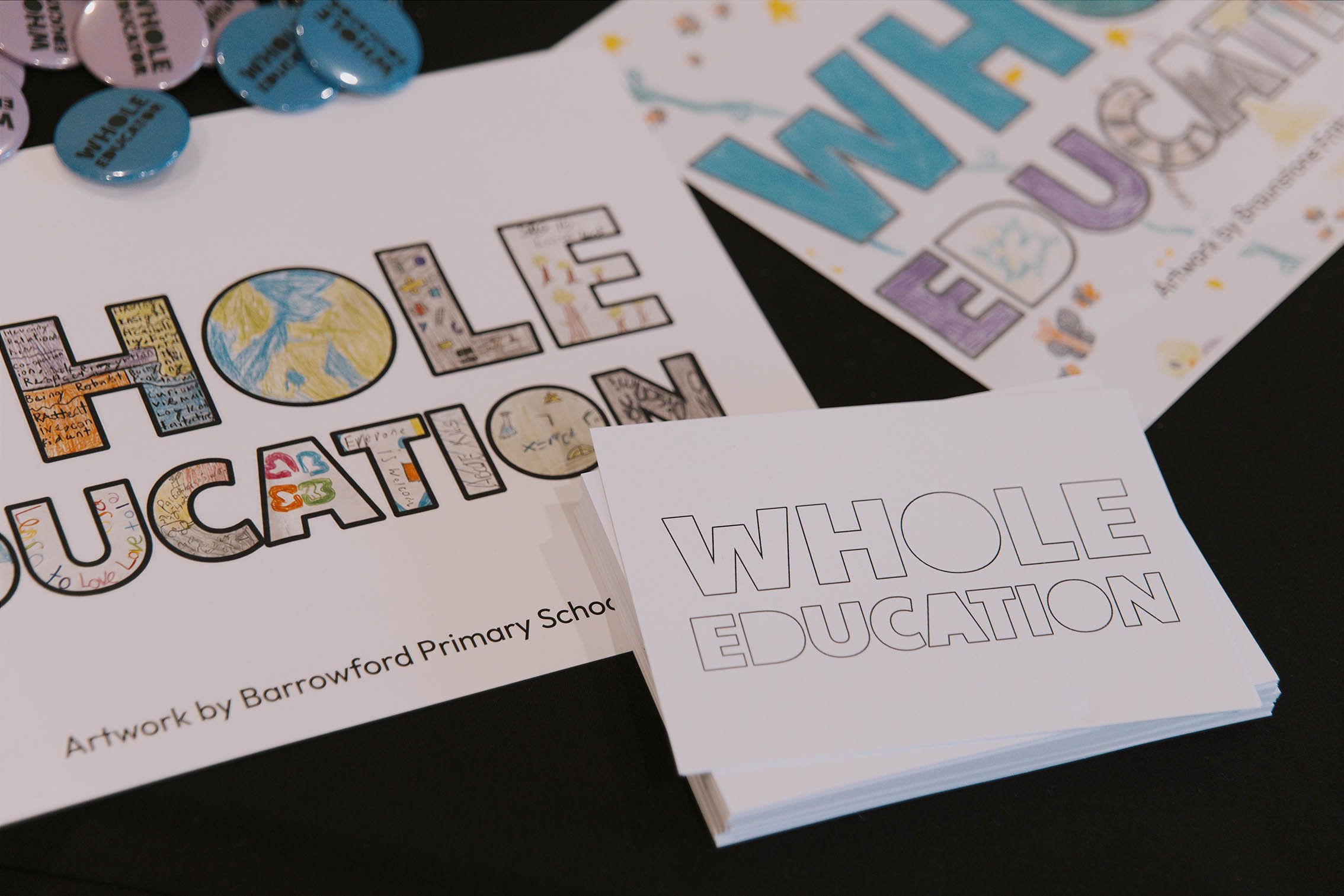 Close-up of printed materials with colorful, hand-drawn style lettering and illustrations. The text on one paper reads "LOVE WHERE YOU EDUCATION" with the words filled with drawings and colors. Another paper partially visible says "WHOLE" in large outlined letters. There are also round, blue, and white buttons or badges with text on them, and some scattered small illustrations on the papers. The papers are laid on a dark surface.