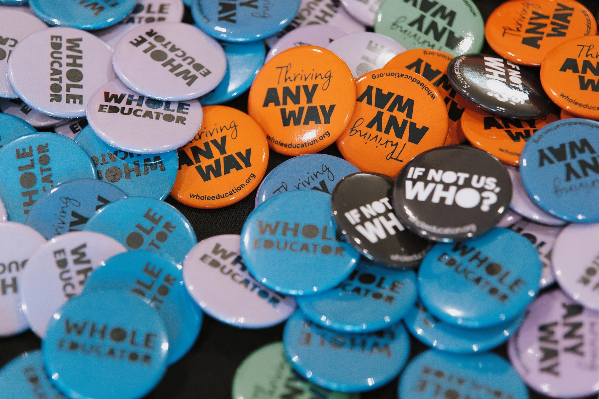 A collection of colorful buttons with political messages, including phrases like 'Whole Educator,' 'Thriving Any Way,' and 'If Not Us, Who?' on a black background.