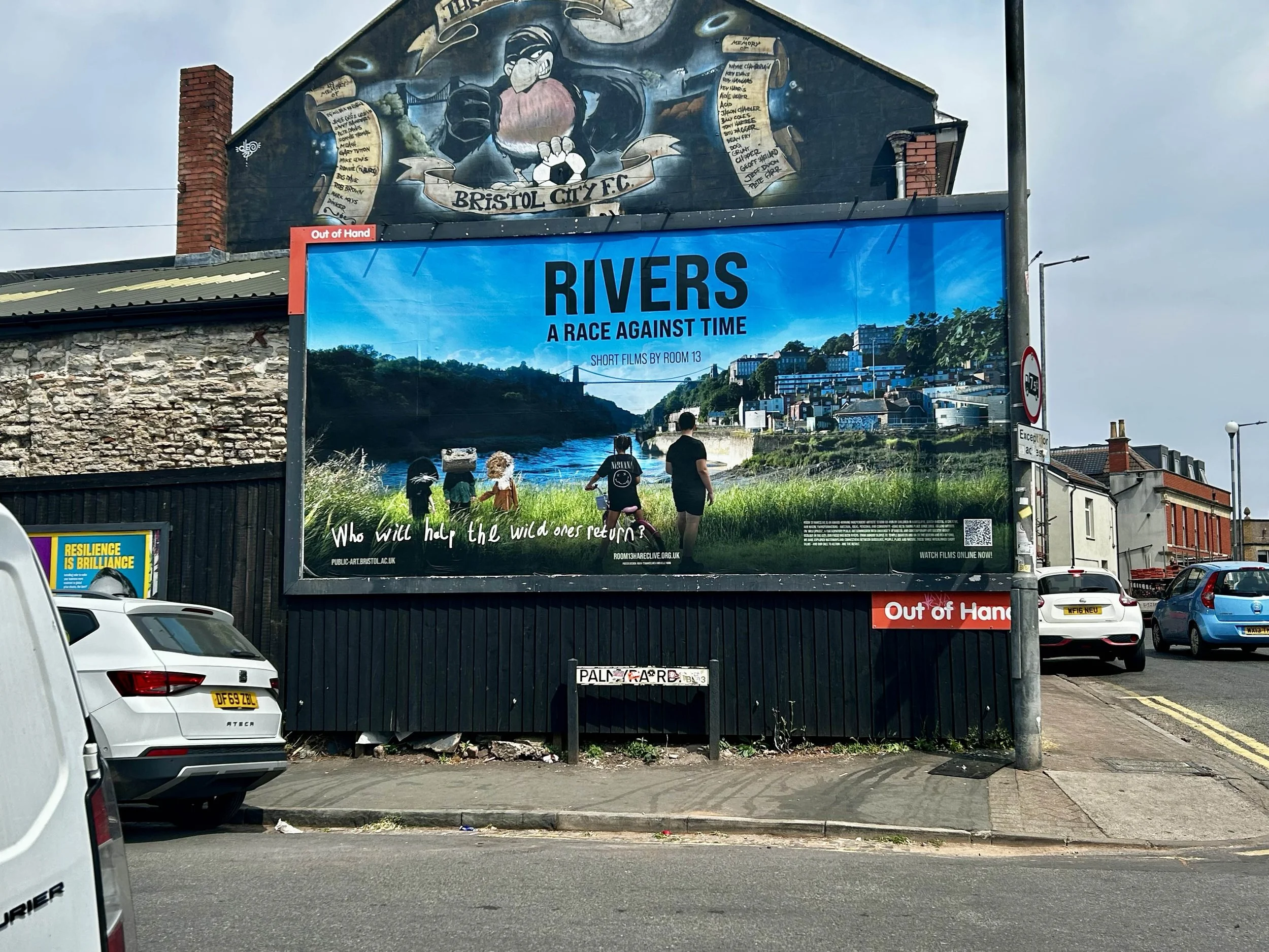 A large billboard featuring Bristol City F.C. and a mural of a soccer player, with a river and cityscape in the background, and people walking along the pathway in front of it.