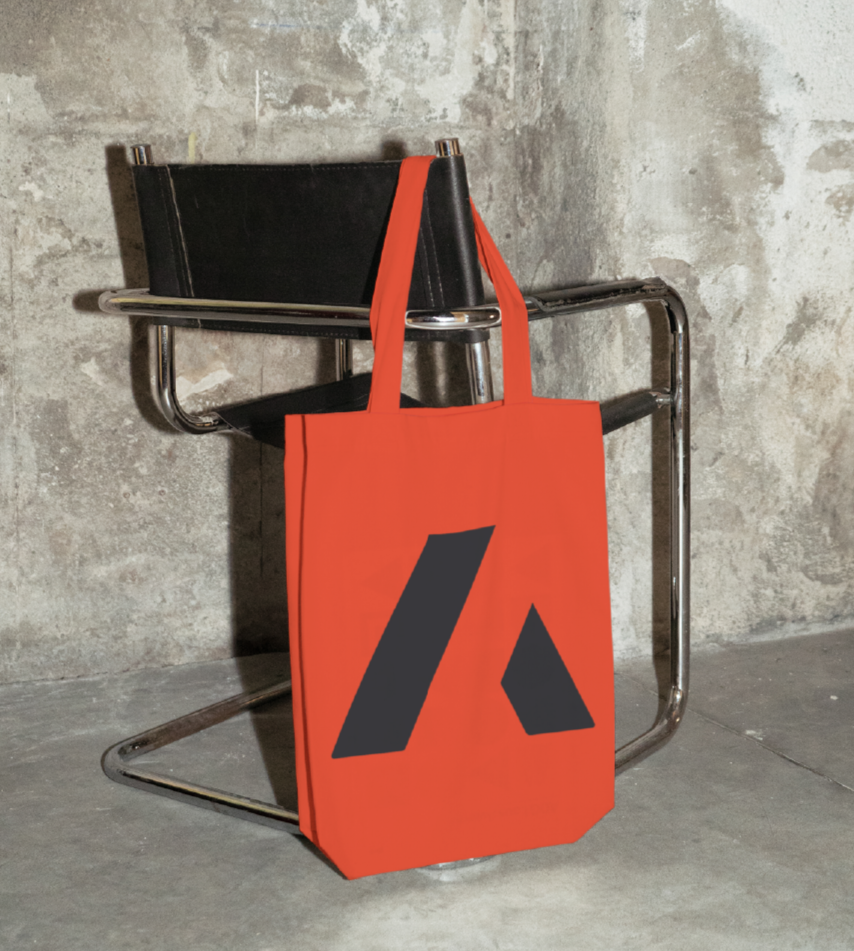 Orange tote bag with black logo hanging on a chair against a textured concrete wall.