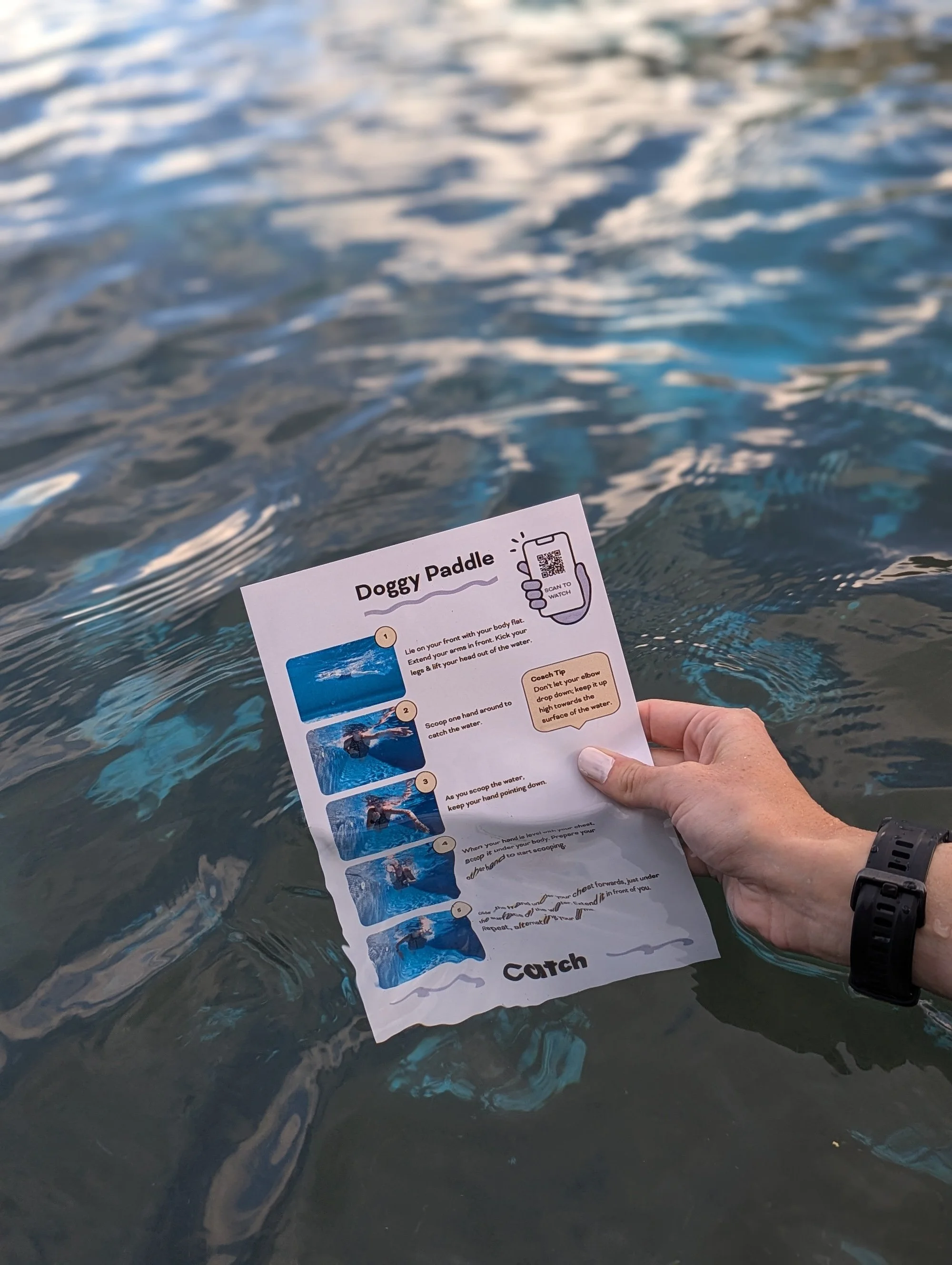 Person holding a sheet of paper titled 'Doggy Paddle' with instructions, partially submerged in water. The water is blue with visible ripples, and the person's hand is near the bottom right corner, wearing a black watch.