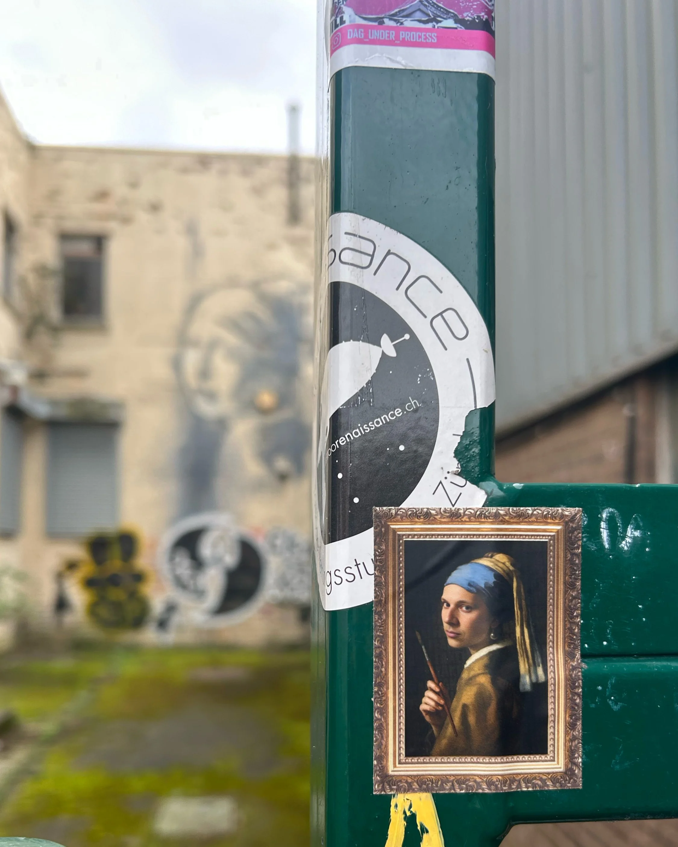 Close-up of graffiti on a green pole, with a painting of a person in historical attire framed like a portrait. The person holds a thin stick or pointer and has a serious expression.