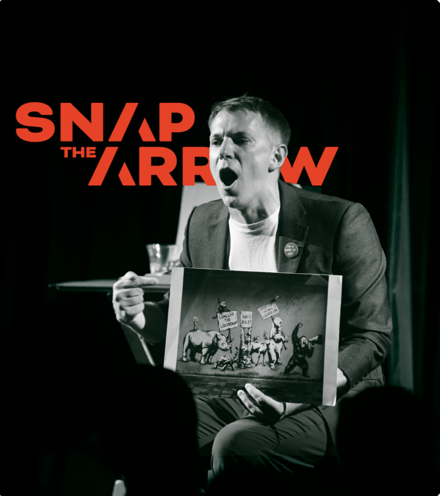 A man in a suit holding a framed picture, speaking passionately during a performance or presentation titled 'SNAP THE ARROW' with a dark background.