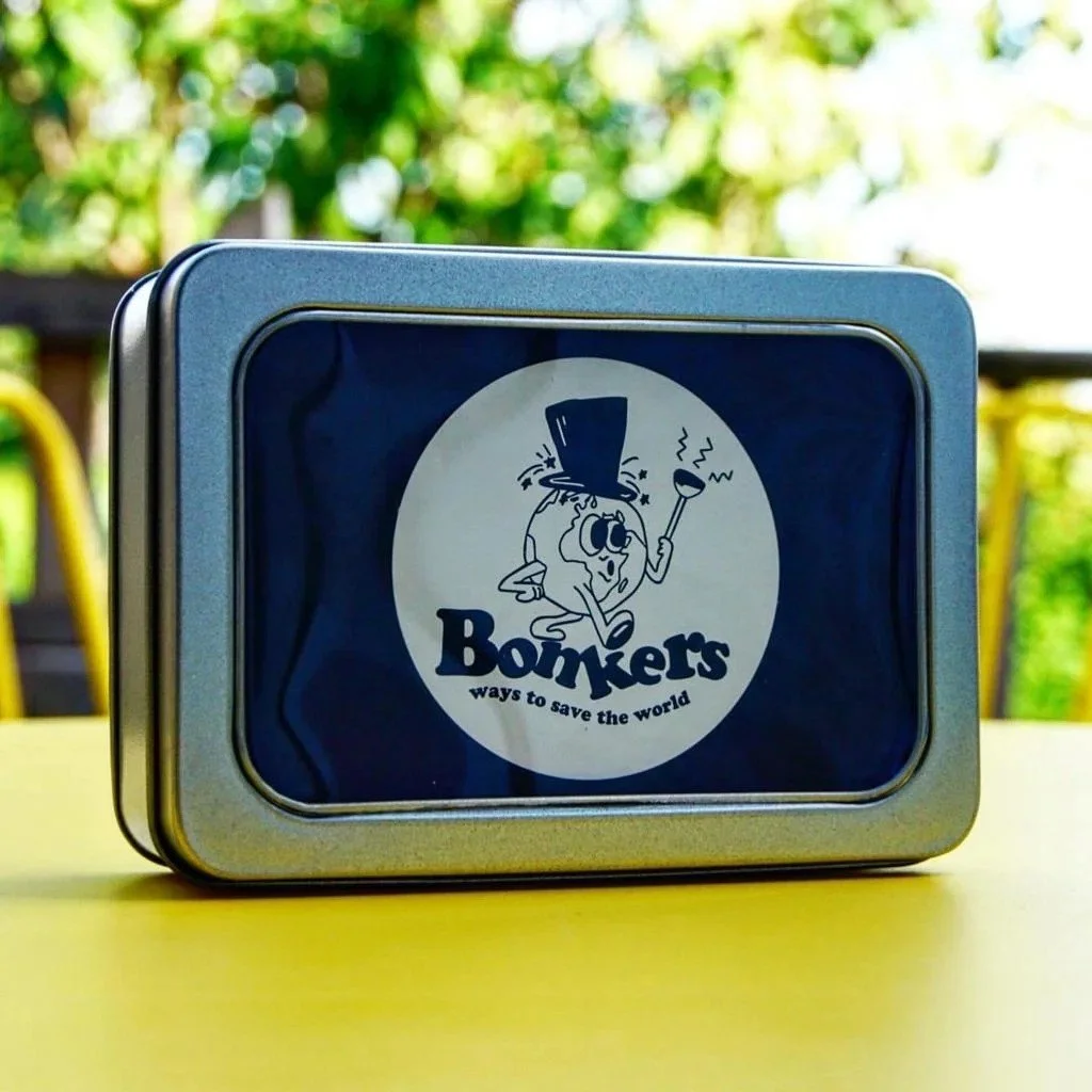A metal tin box with a cartoon character of a dog wearing a top hat and holding a smoking pipe, with the text "Bonkers ways to save the world" on the label. The background shows trees and a yellow surface.