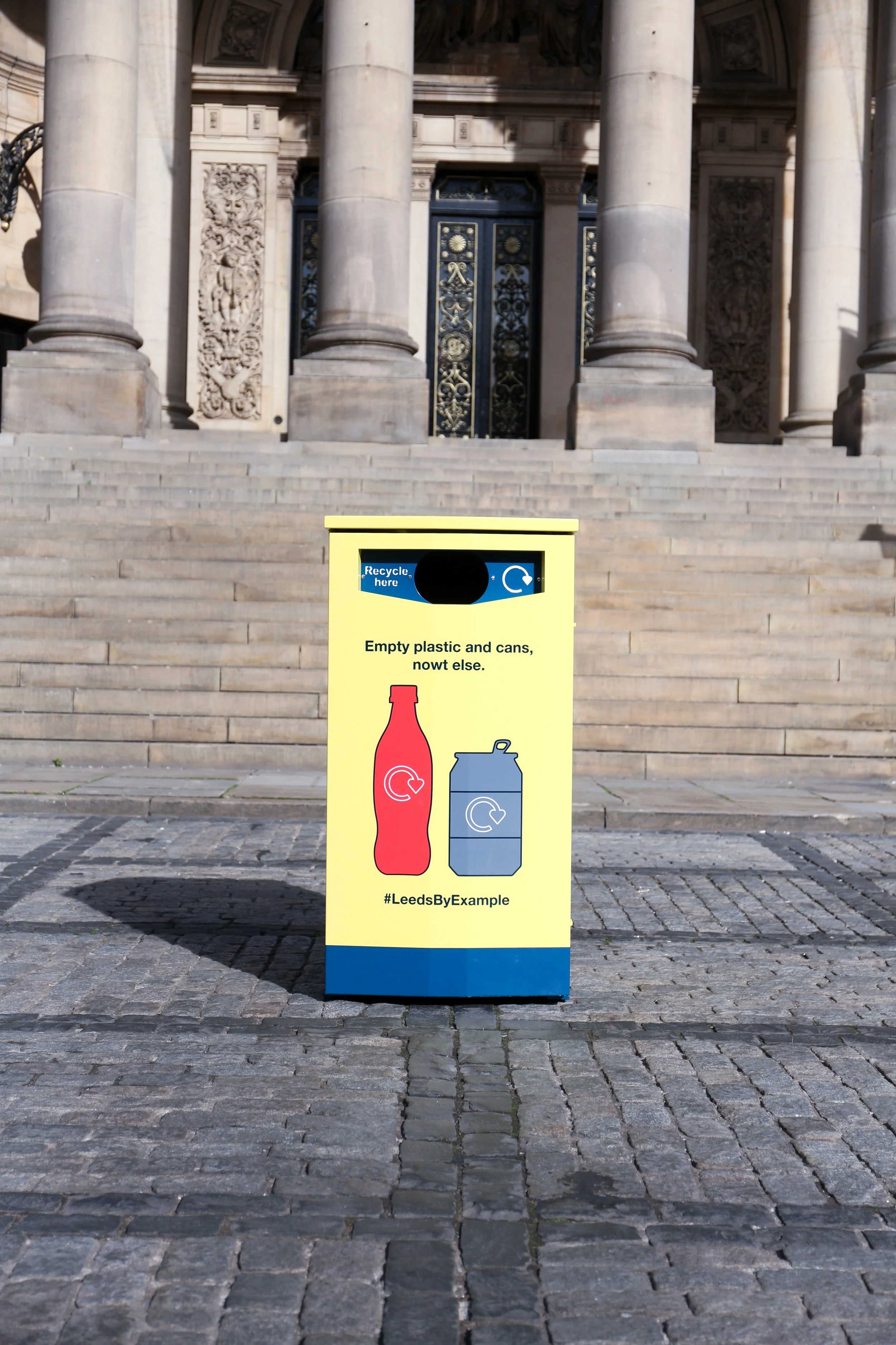 Yellow recycling bin with a graphic of a plastic bottle and a can, set against stone steps in front of a building with columns and ornate details.
