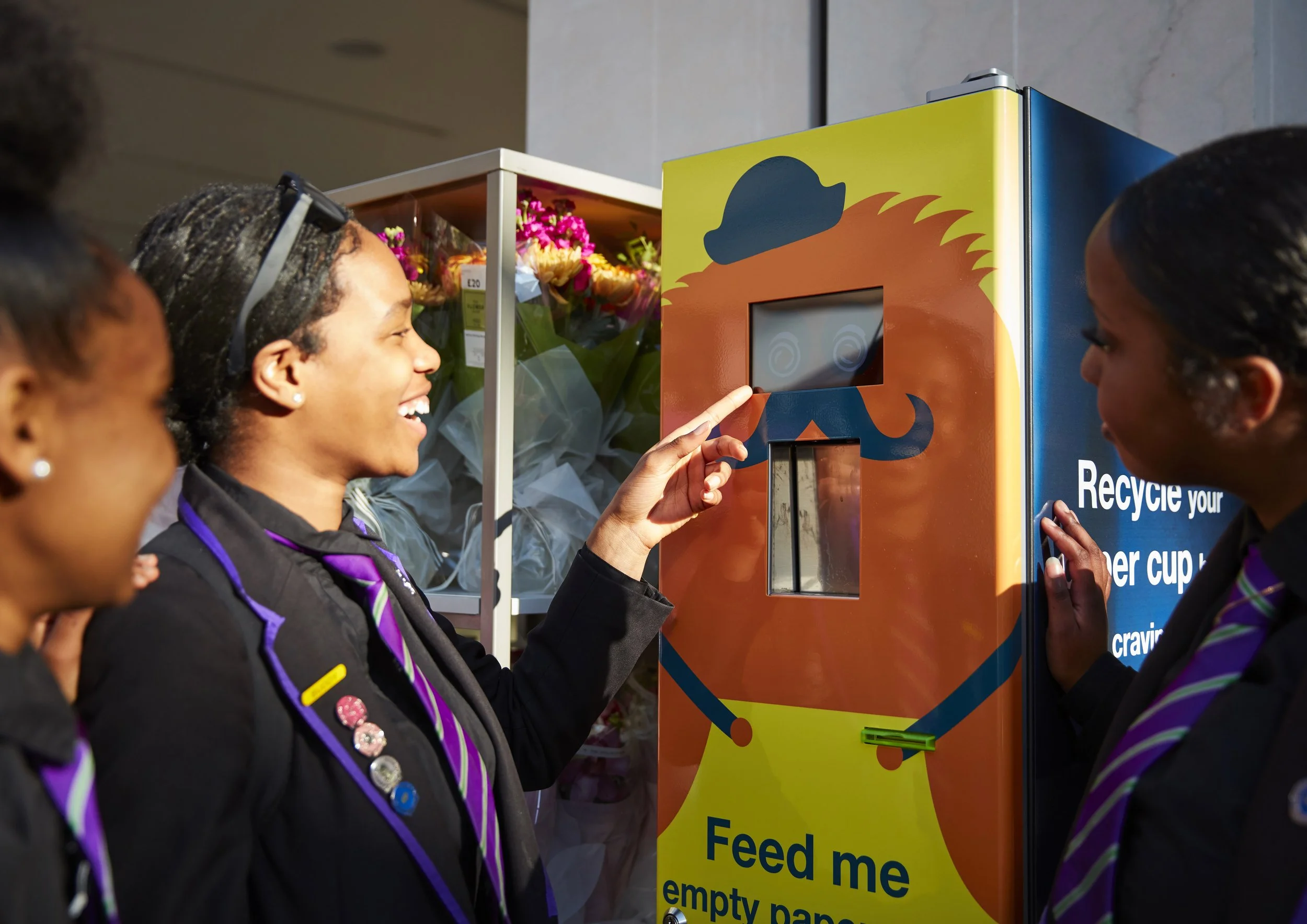 A group of people in uniform using a smart recycling bin in a public space. The bin has a colorful design and a screen for interaction, with text encouraging recycling and feeding the bin.