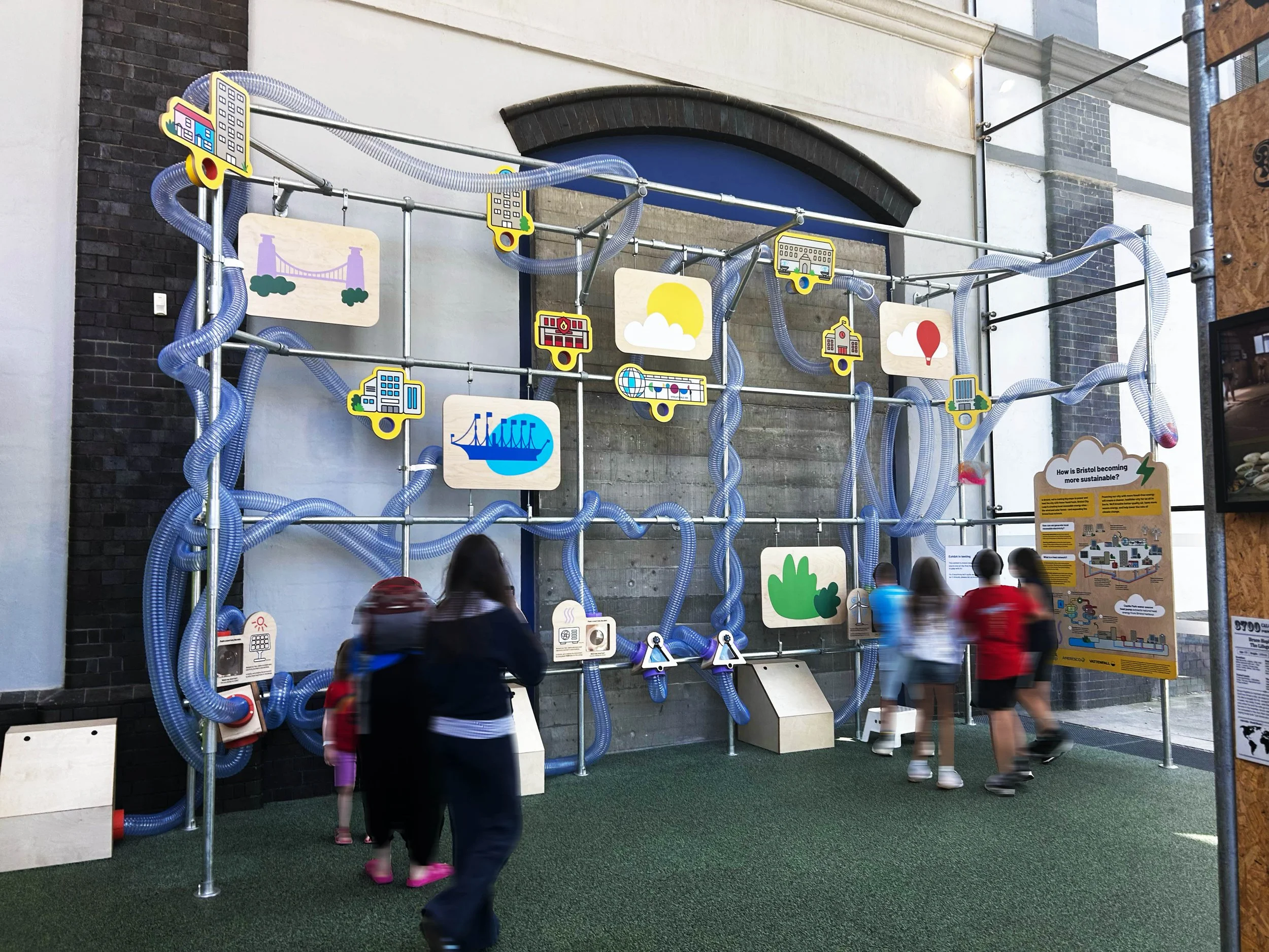 Indoor exhibit with colorful diagrams and illustrations about urban sustainability, including images of buildings, a bridge, nature, and a boat, with people observing.