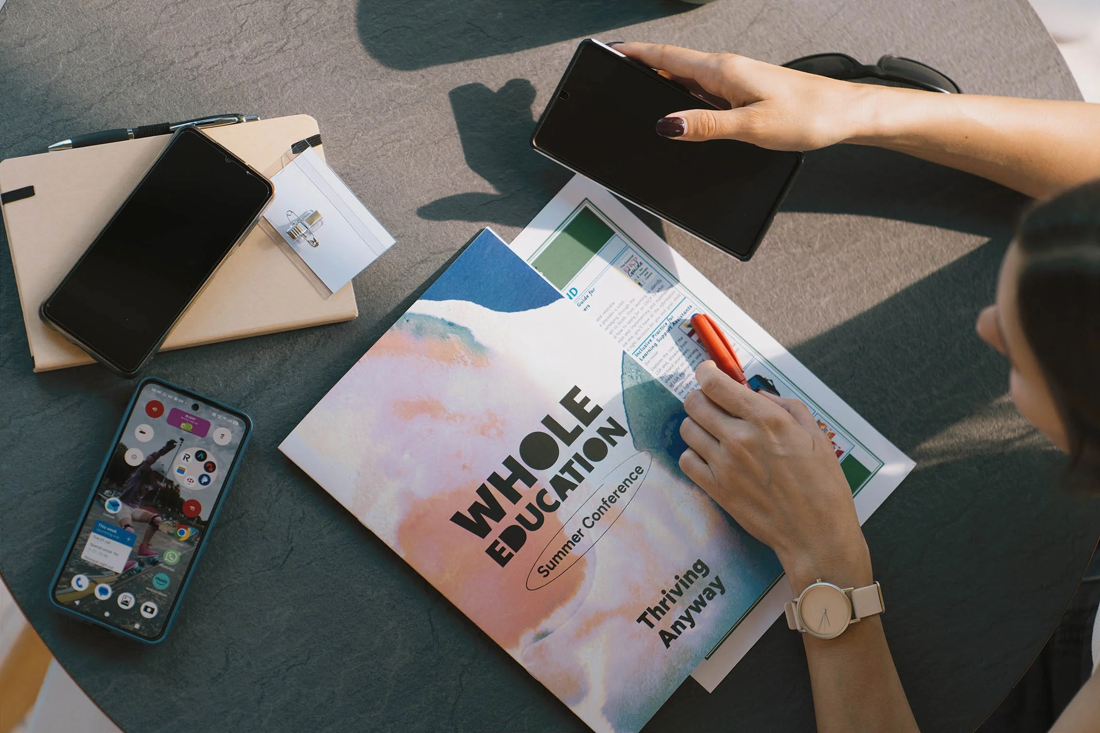A person's hand with dark nail polish holding a smartphone over a table with notebooks, brochures, a palm tree-themed phone case, and a pen. The person's wrist has a beige watch, and their face is partially visible.