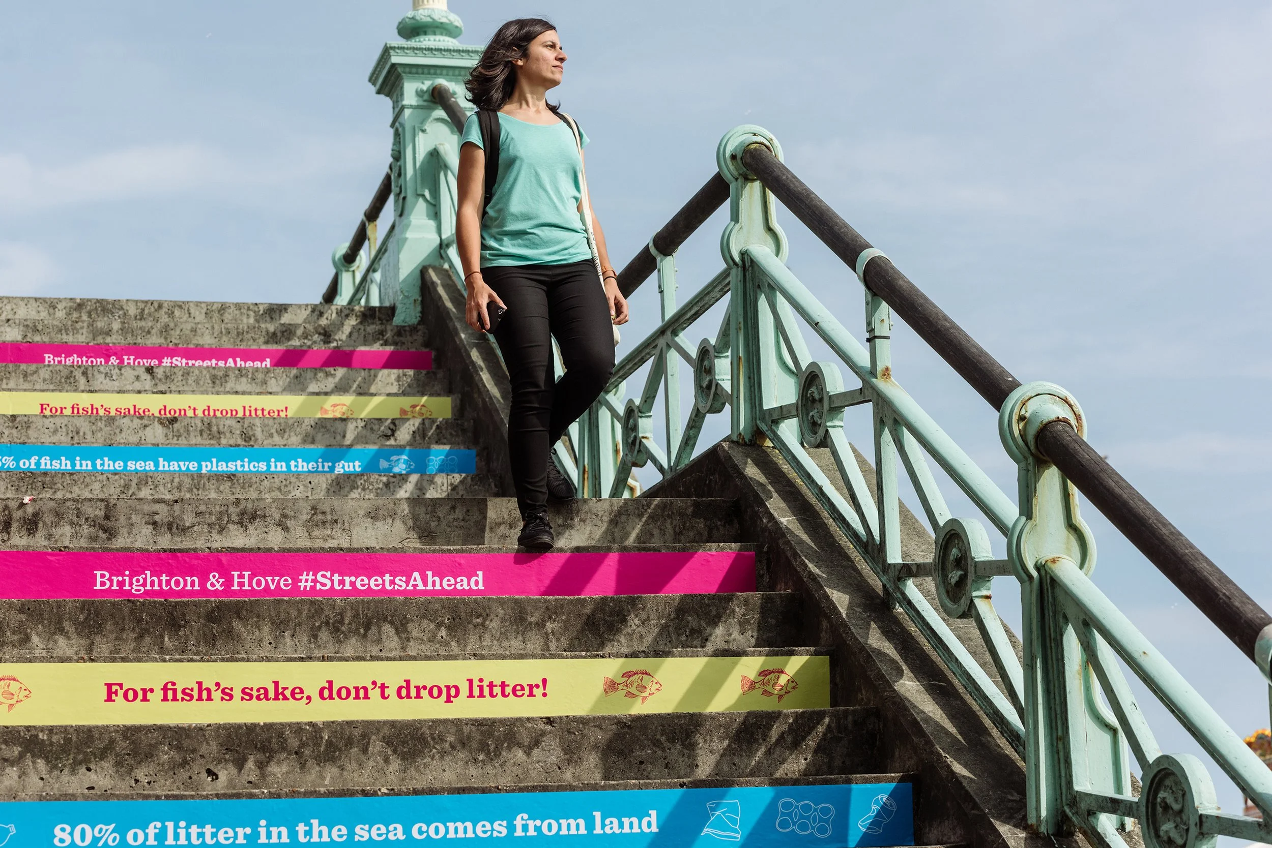 A woman with dark hair, wearing a teal T-shirt and black pants, standing on a set of outdoor stairs next to a teal-colored metal railing, looking into the distance. There are informational and environmental slogans on the stairs about littering and plastic pollution in the sea.