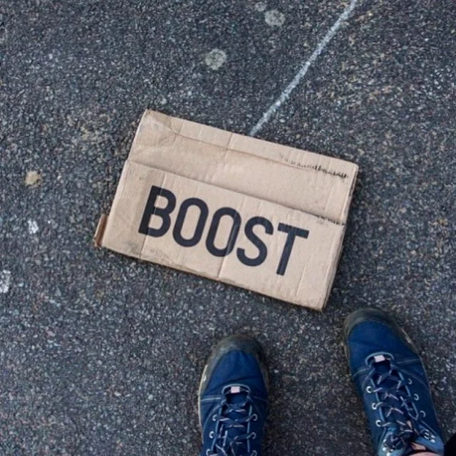 A cardboard box labeled "BOOST" on a rough asphalt surface, with a person wearing blue athletic shoes standing nearby.