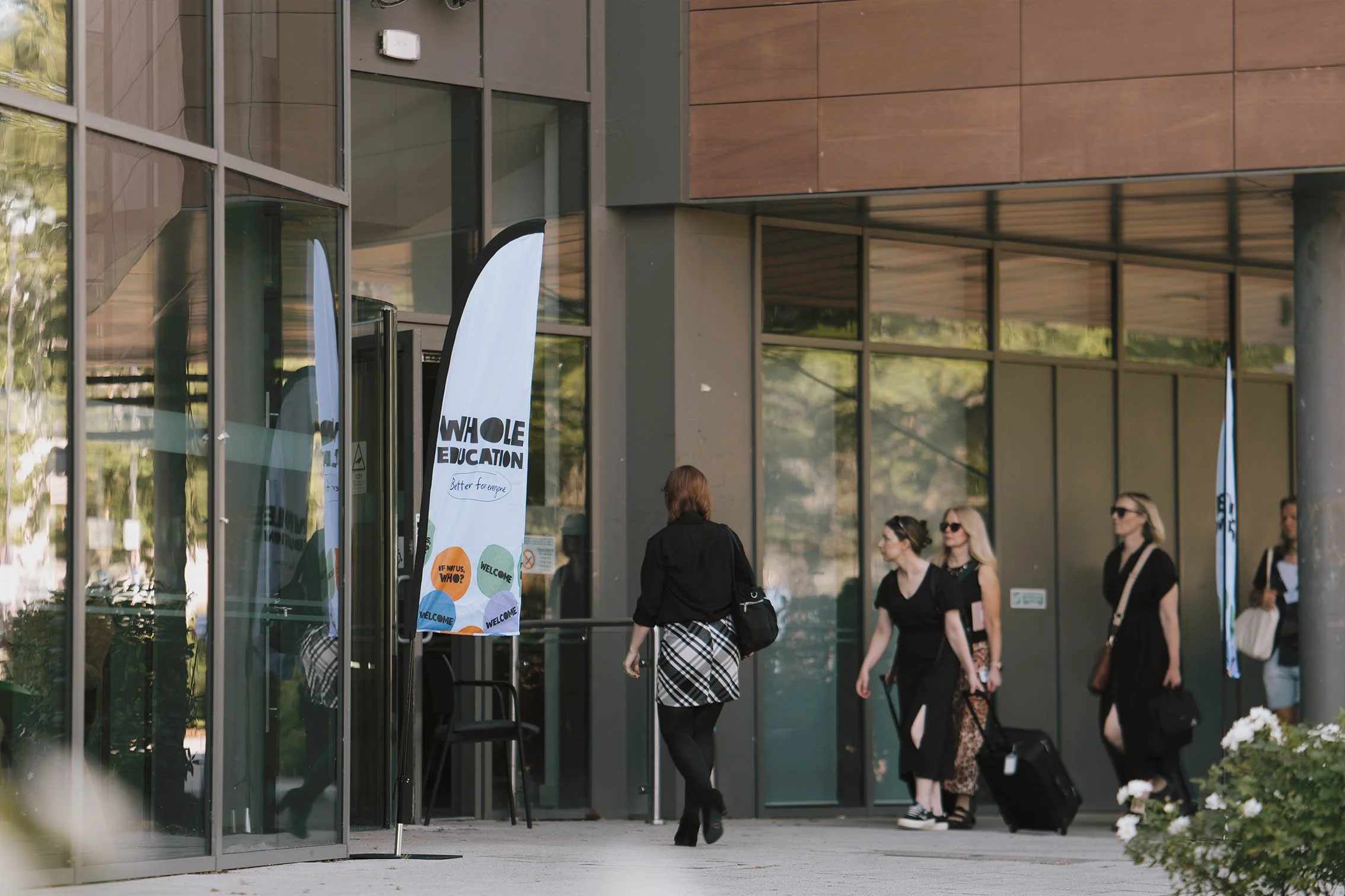 People walking in and out of a modern building with glass doors, some pulling suitcases, and banners outside advertising an event.