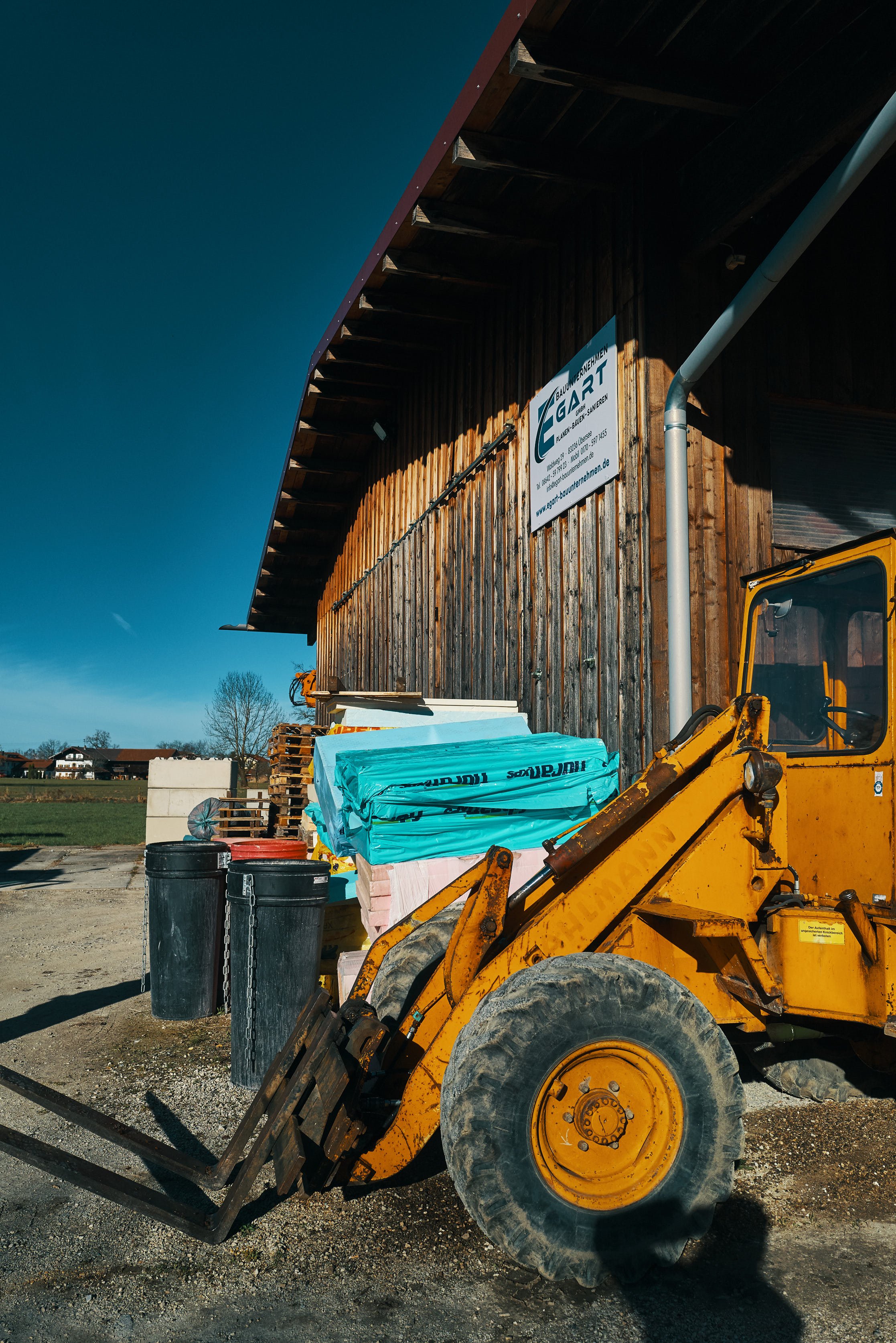 EGART Baustelle mit gelbem Baufahrzeug, Bauzäunen und Holzhaus im Hintergrund, daneben Mülltonnen und Baum.