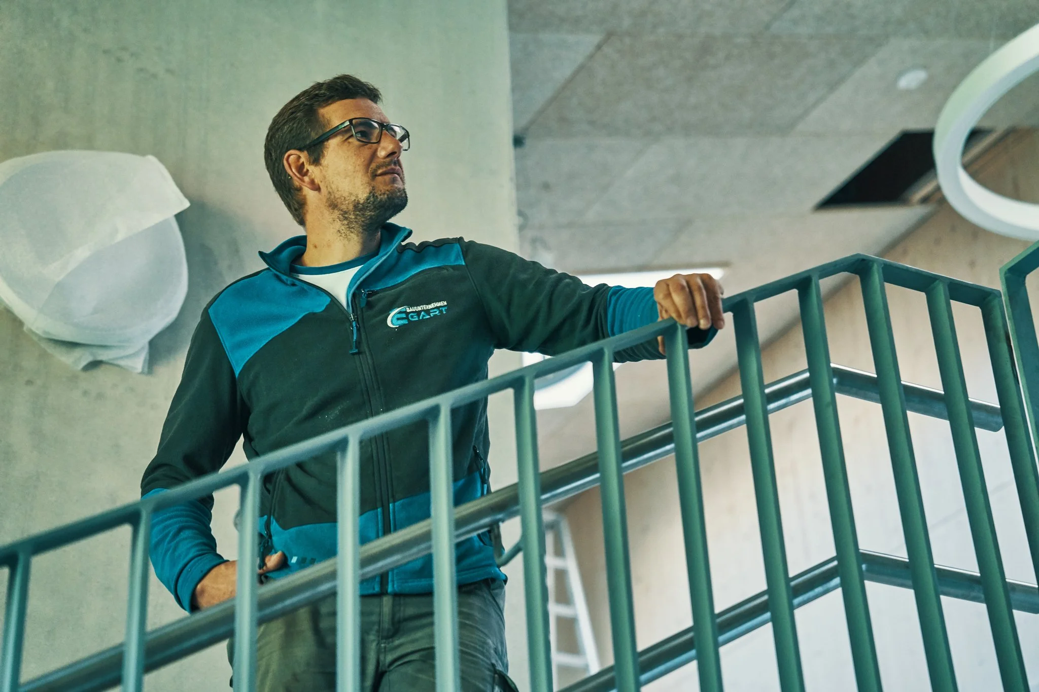 EGART Mitarbeiter mit Brille in schwarz-blauer Jacke steht am Geländer eines modernen Gebäudes mit grauer Betonwand.
