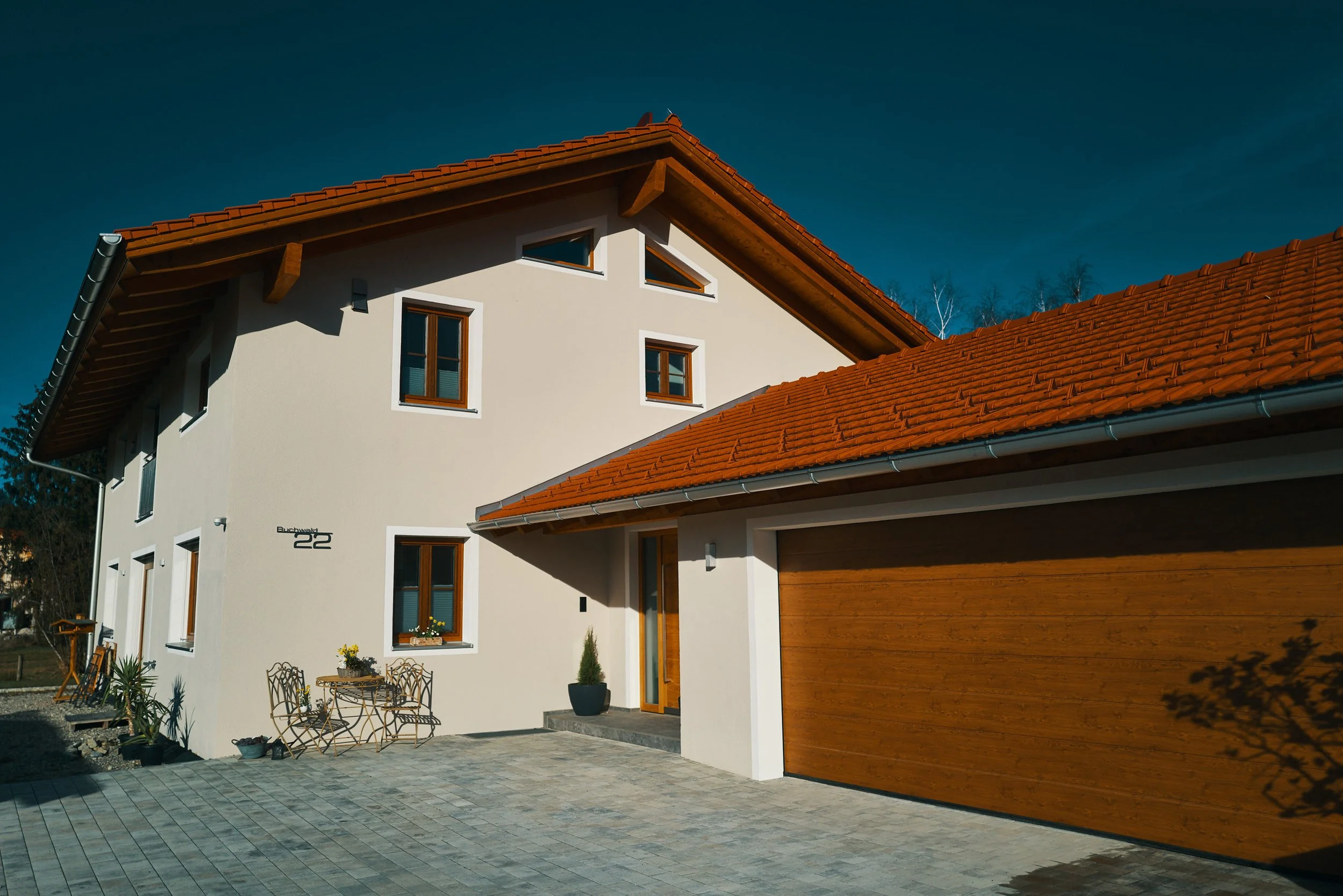 EGART Einfamilienhaus mit Ziegeldach, weißen Wänden und Holztür, gepflasterter Hof mit Gartenmöbeln und Pflanzen unter blauem Himmel.