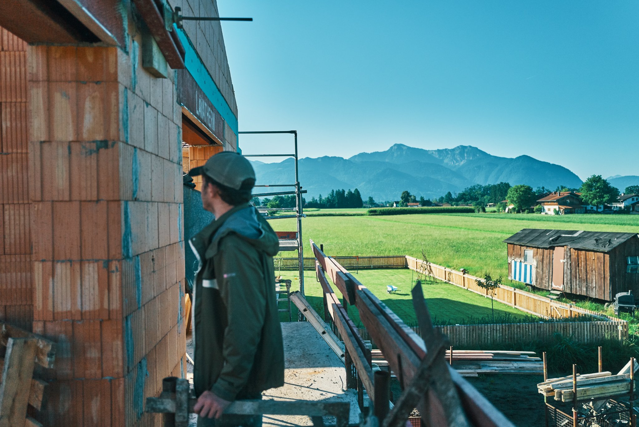 EGART Hausbau auf der Baustelle mit Bauarbeiter, Blick auf Berge und grüne Wiesen bei Sonnenschein.