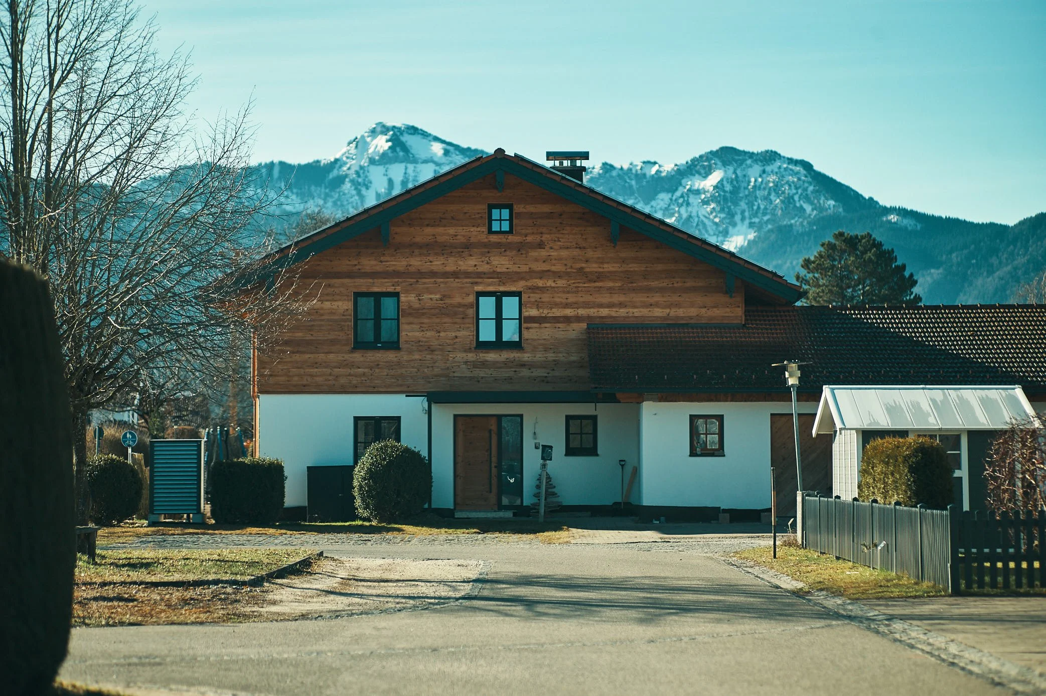 EGART Neubau mit Holzfassade vor Bergkulisse, modernes Haus mit Bäumen ohne Blätter und blauem Himmel.