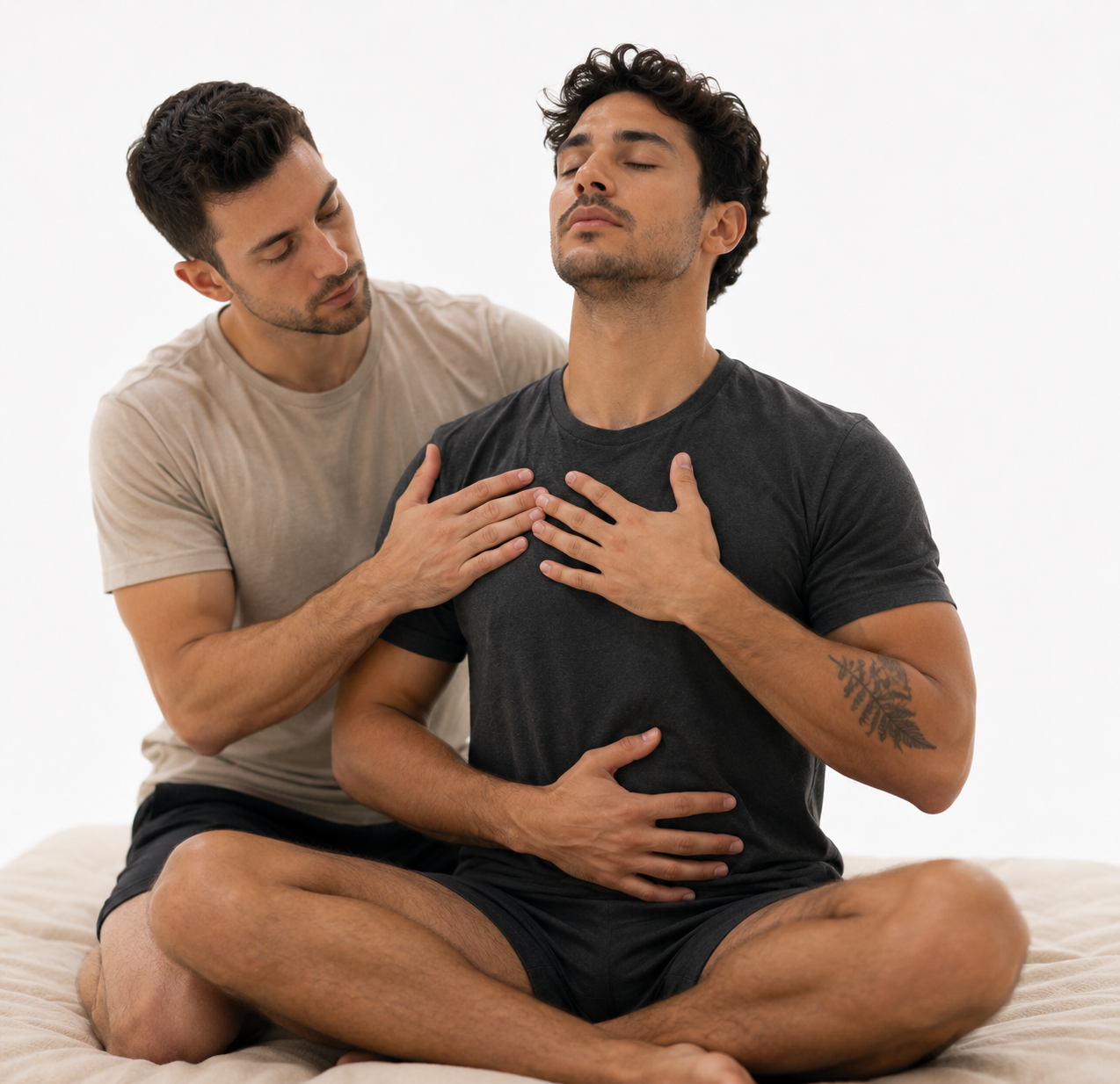 Two men are practicing mindfulness or meditation; one is sitting cross-legged with eyes closed, placing one hand on his chest and the other on his stomach, while the other man is gently touching his shoulder and chest.