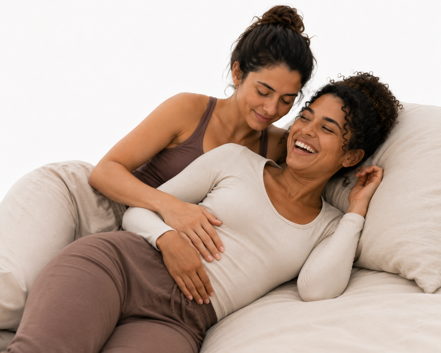 Two women are on a beige sofa, one lying down and the other leaning over her, both smiling and sharing a joyful moment.
