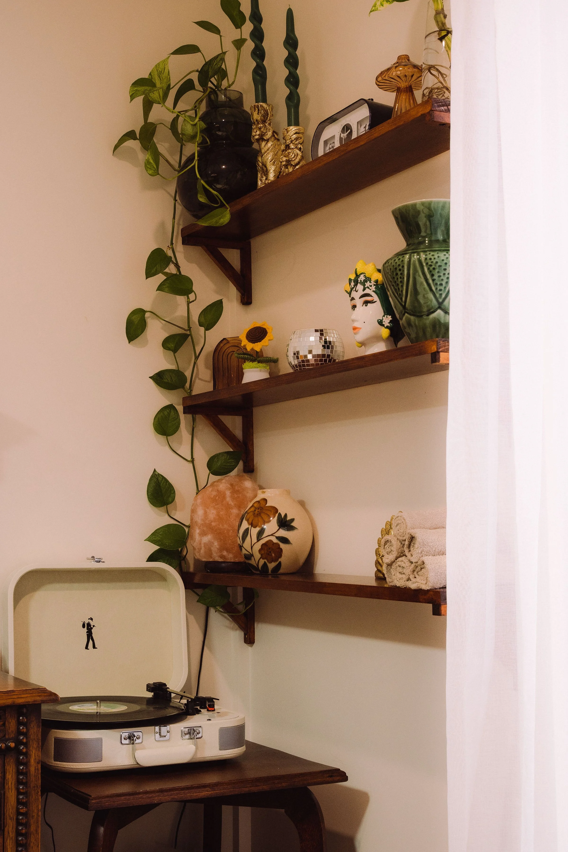 A vintage record player on a wooden table with shelves above holding decorative items, including vases, pottery, a disco ball, a ceramic bust, toy animals, textiles, and a leafy vine plant.